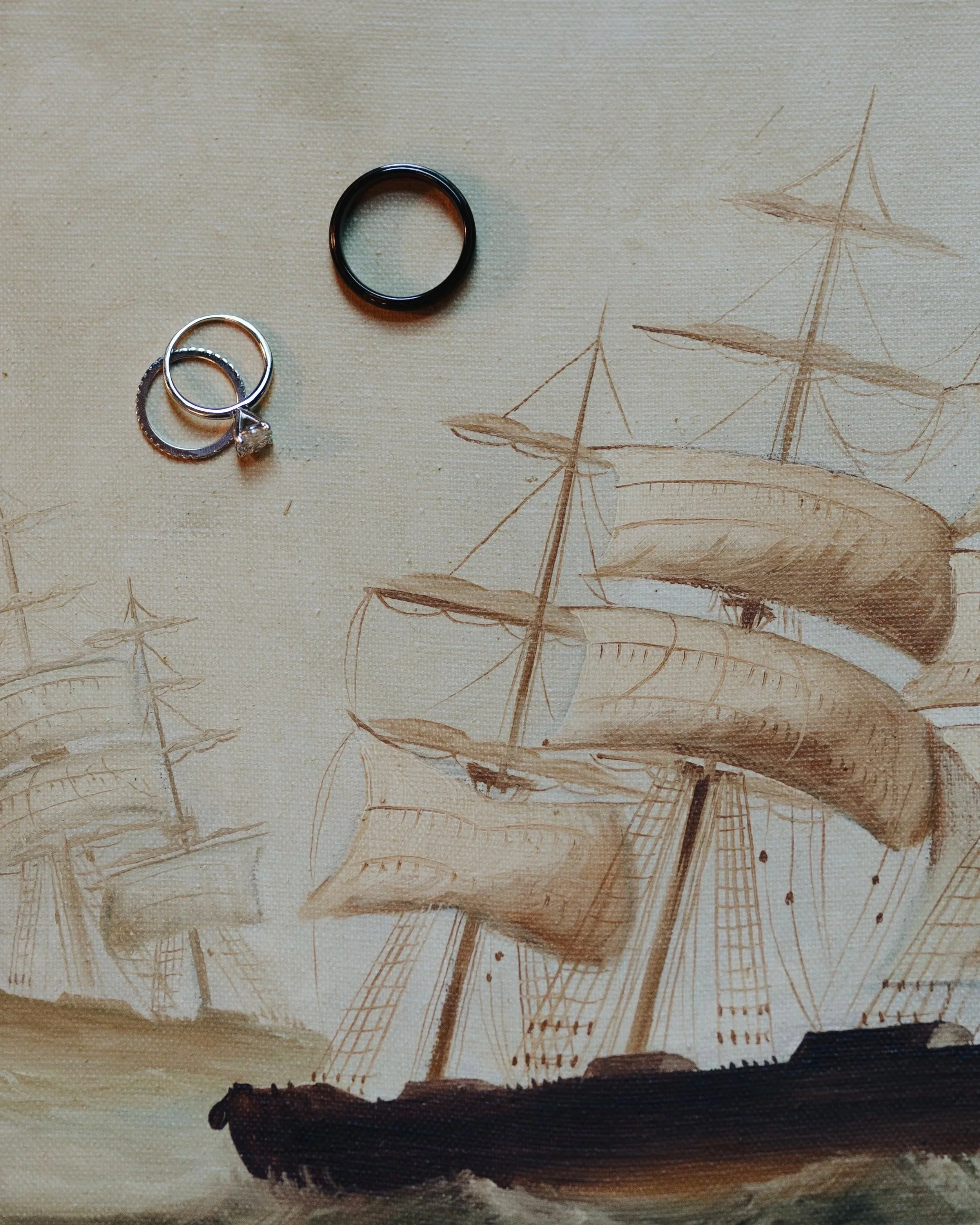 Two rings, one black and one silver with a diamond, placed on a fabric with a painting of sailing ships. DOCUMENTARY STYLE, EDITORIAL STYLE