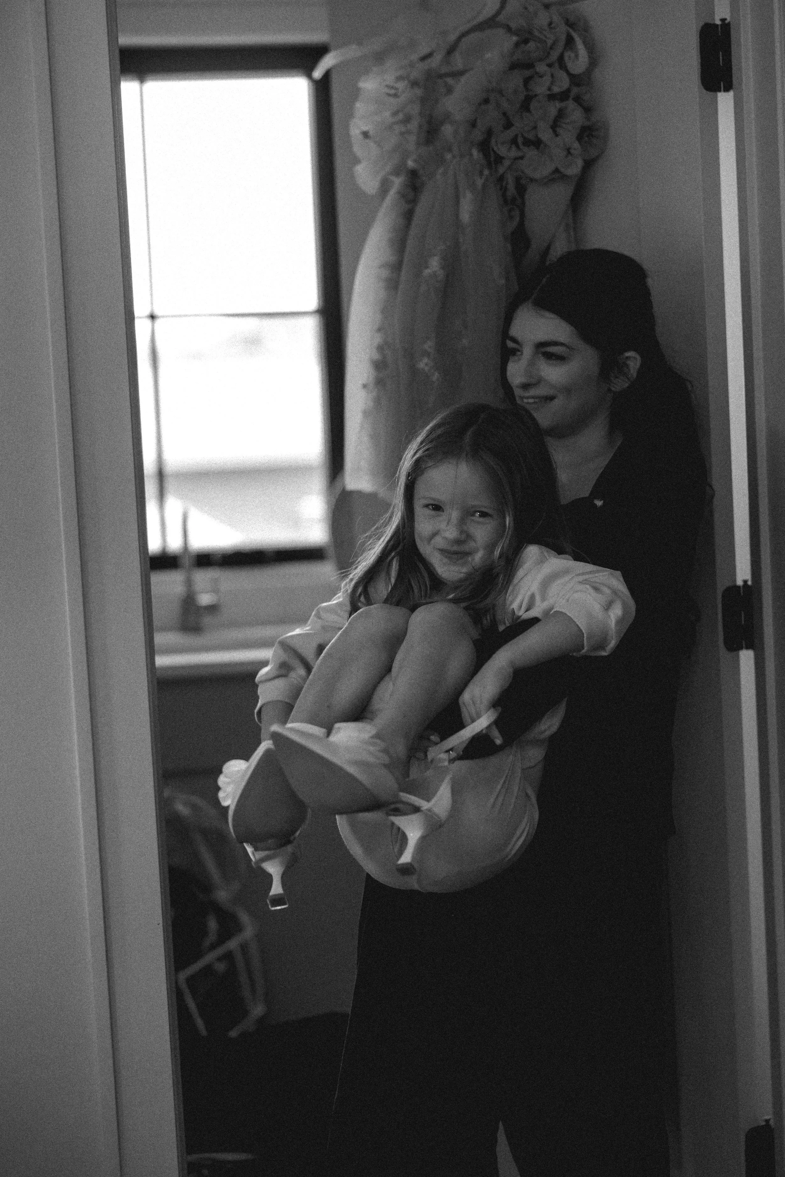 A woman is standing in a doorway, holding a young girl on her lap. The girl looks at the camera with a playful expression. The background shows a window and some clothing hanging. DOCUMENTARY STYLE, EDITORIAL STYLE
