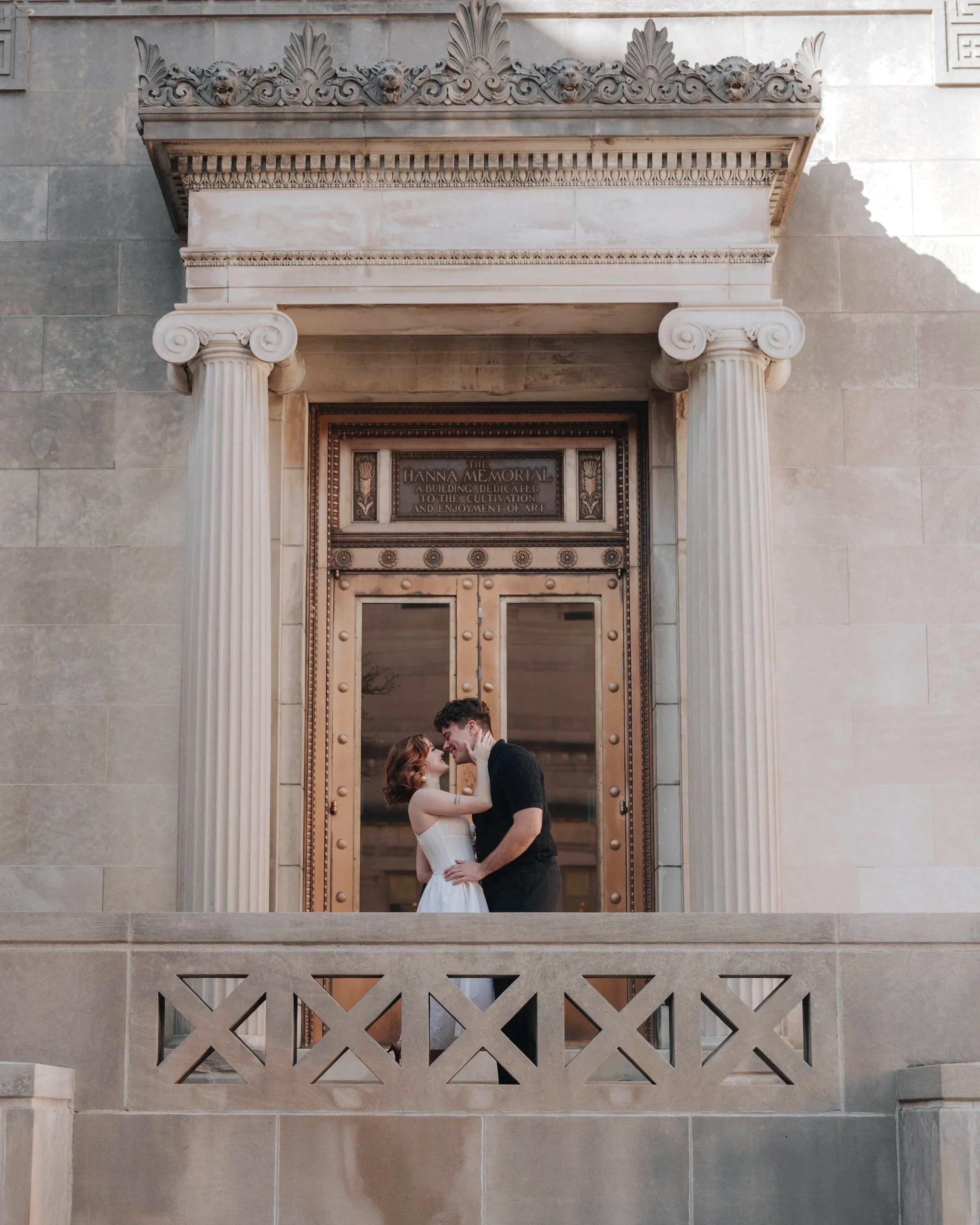A couple standing close together in front of a historic building with ornate columns and decorative carvings, embracing and touching foreheads.