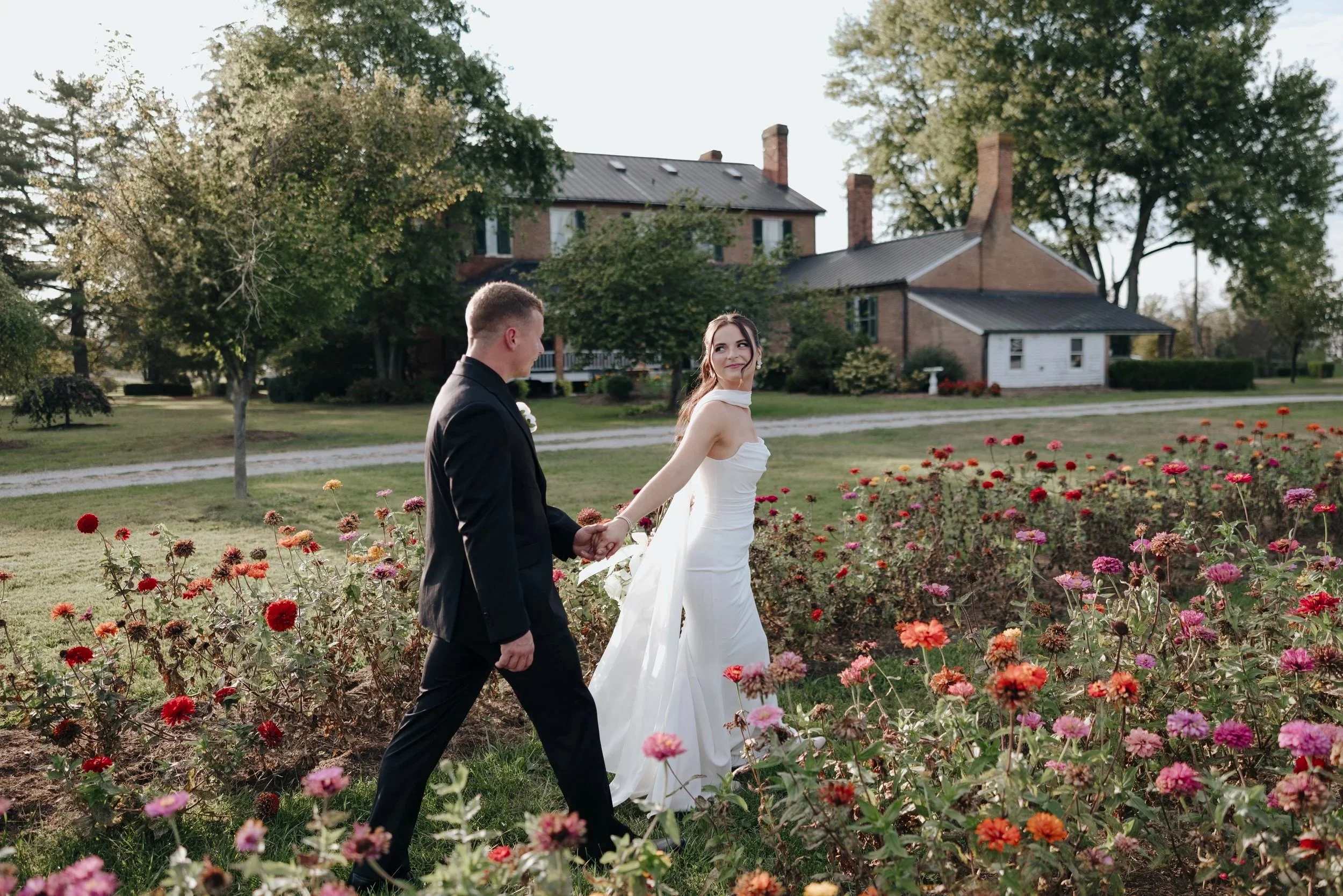 A bride and groom holding hands and walking through a flower garden with a house and trees in the background during daytime.