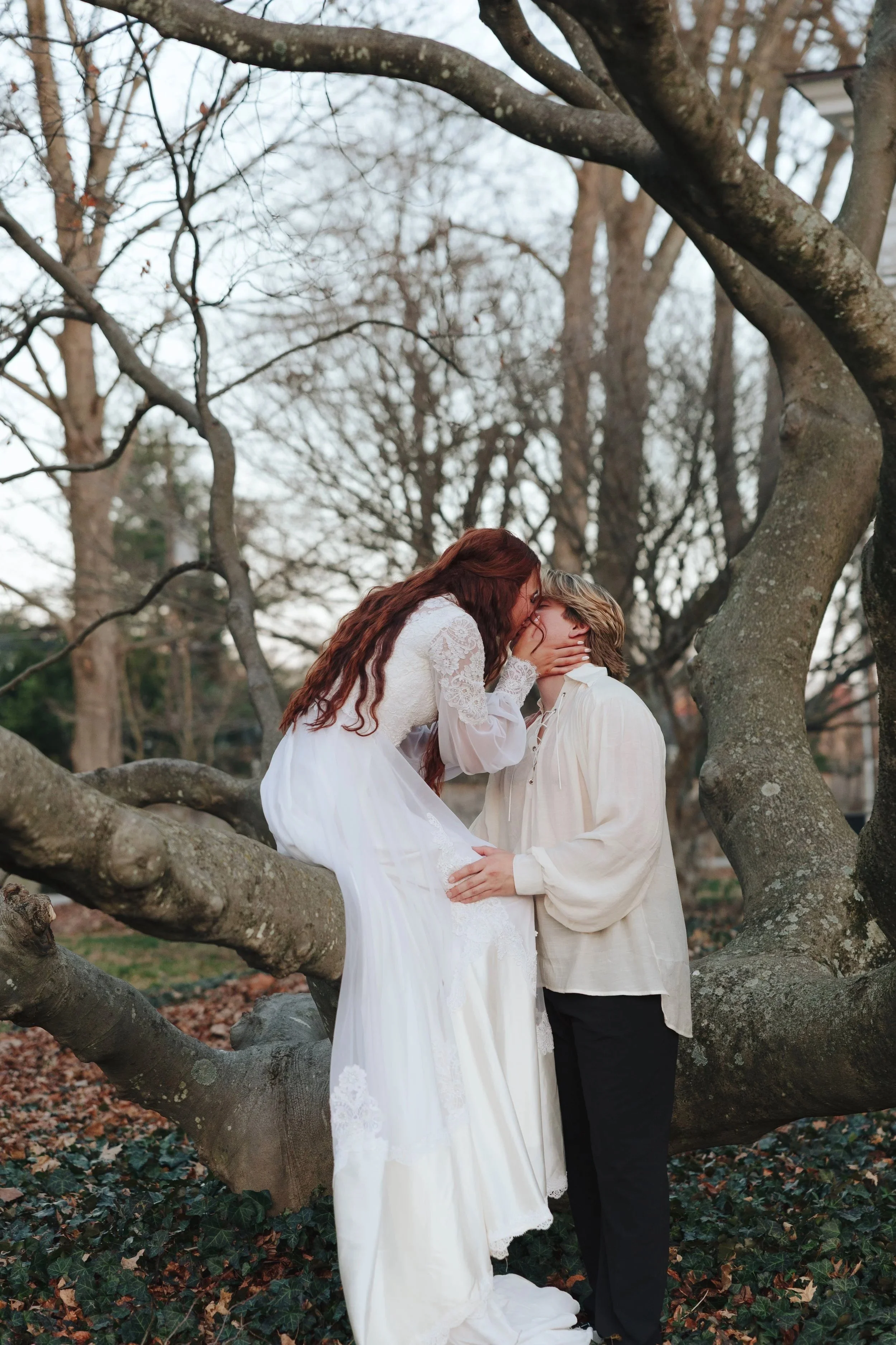 A couple sharing a kiss outdoors during fall, with the woman sitting on a large tree branch and wearing a white dress, and the man standing and holding her.