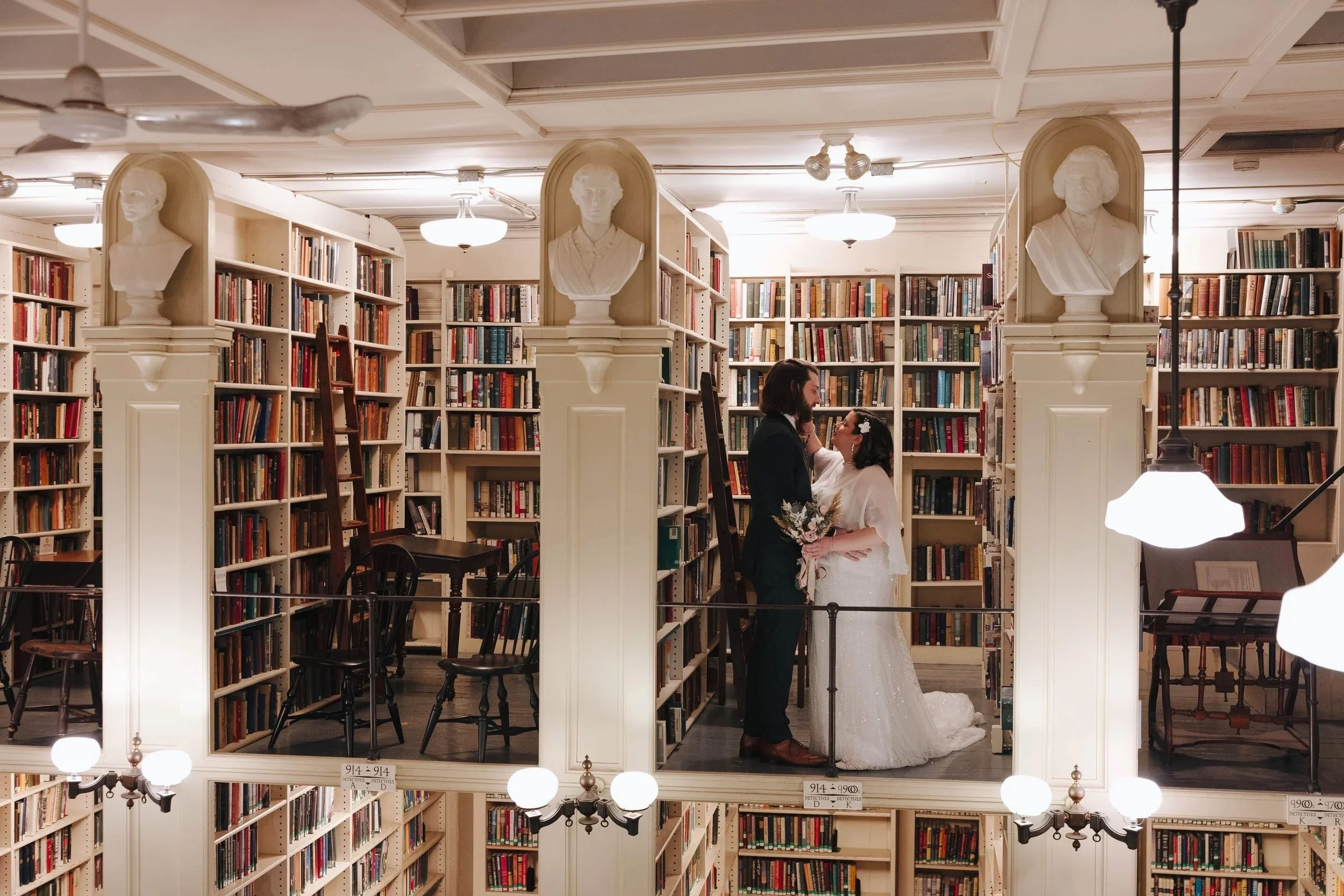 A couple in wedding attire standing close in a library, with bookcases filled with books behind them. The bride is holding a bouquet and wearing a white wedding dress, while the groom is dressed in a dark suit. The scene is reflected in a mirror on t