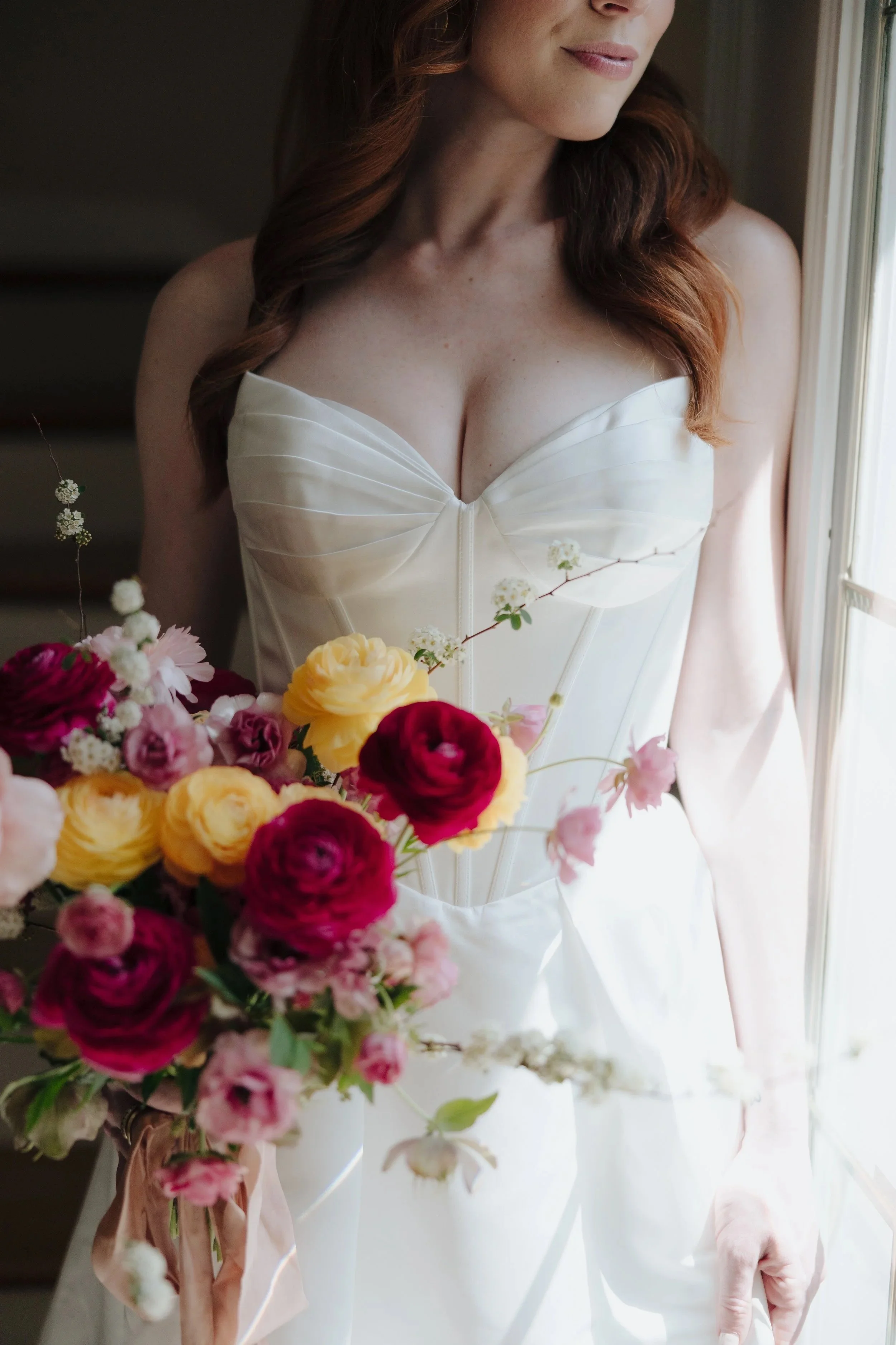 Woman in a white strapless dress holding a bouquet of pink, yellow, red, and white flowers, standing near a window.