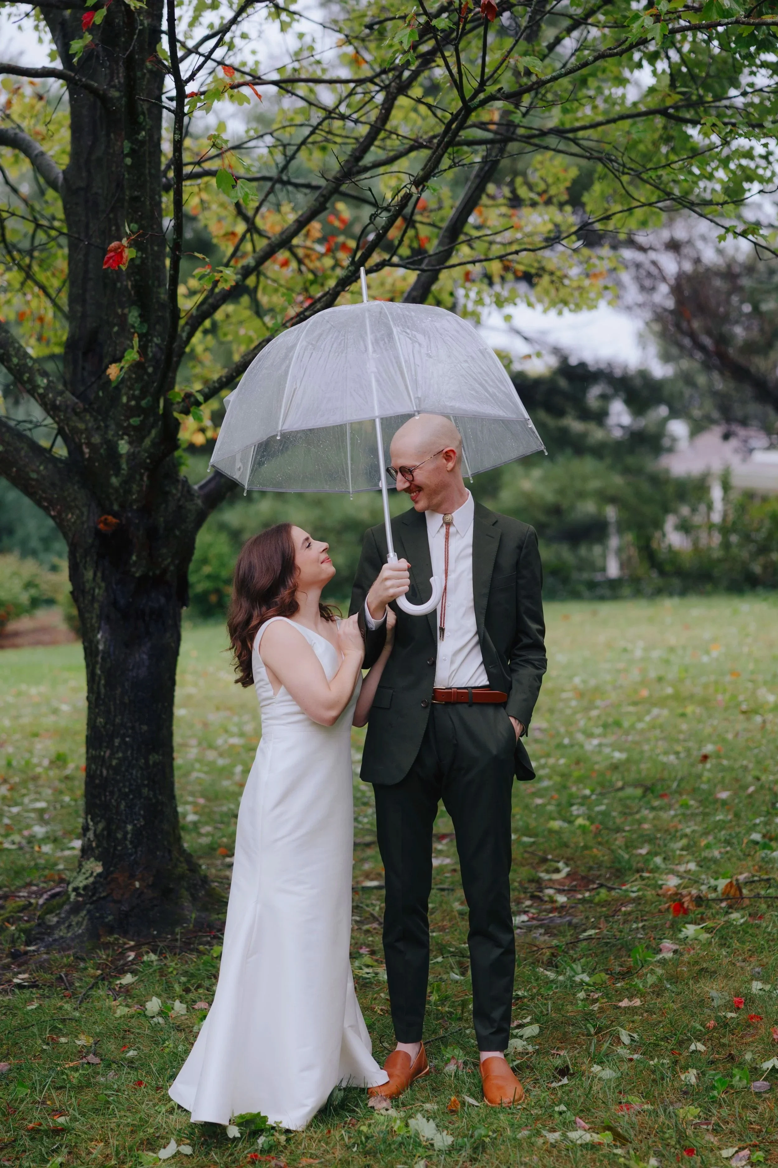 A woman in a white dress and a man in a dark suit and glasses stand outside under a large transparent umbrella during rainy weather. They are smiling and looking at each other near a tree with autumn leaves.