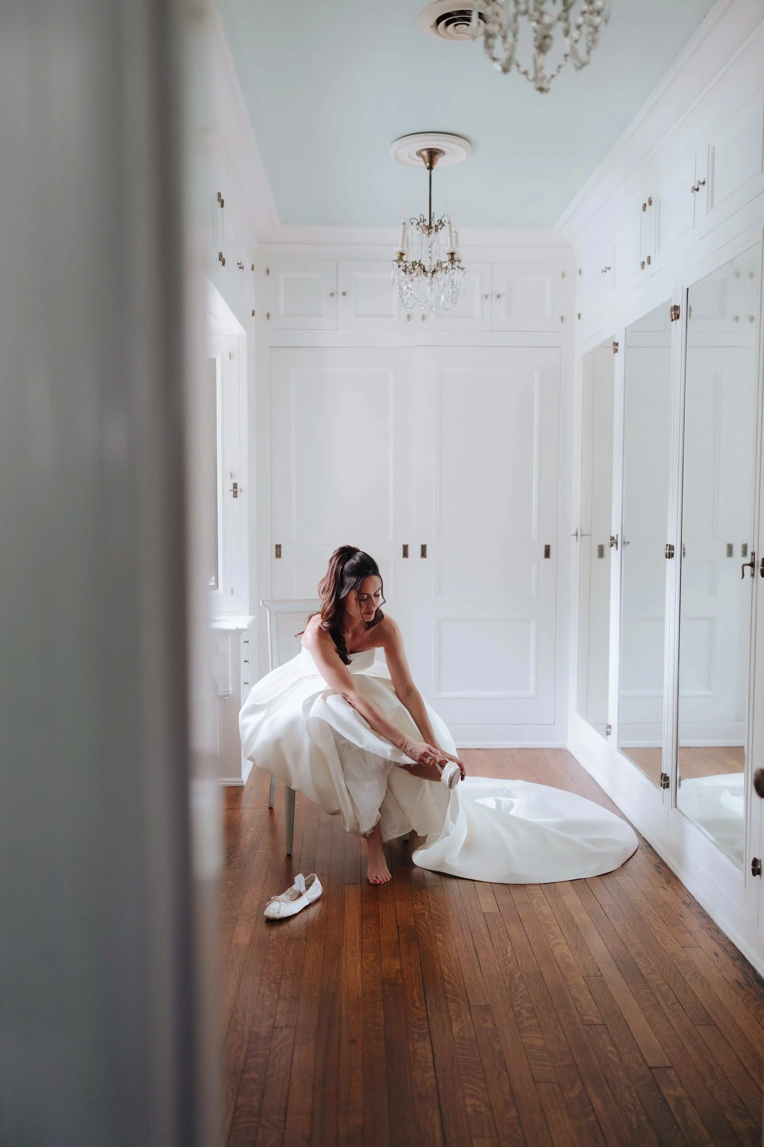 A woman in a wedding dress is sitting on a chair in a white room, putting on her shoes. The room has white paneled walls, polished wooden floors, a chandelier, and mirrored closet doors. There are a pair of white shoes on the floor in front of her.