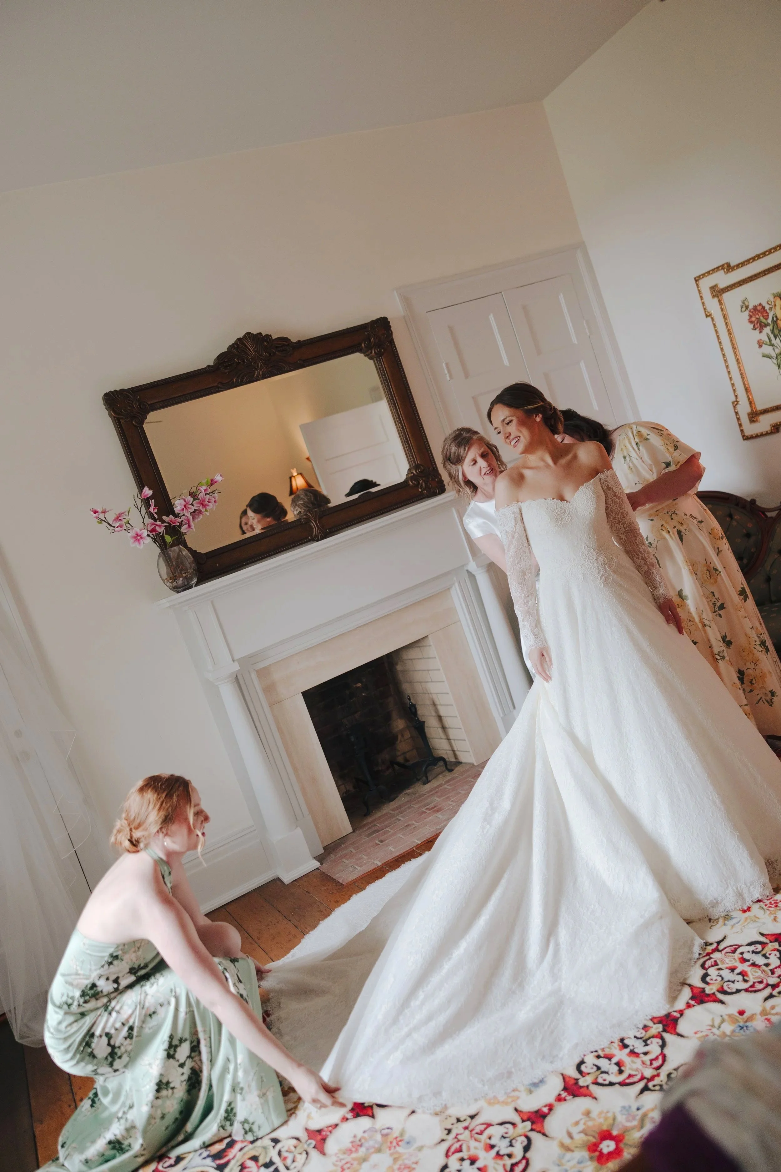 A bride in a white wedding dress is being assisted by several women, including one in a floral dress and another in a white satin blouse, as they prepare. The scene takes place in a living room with a fireplace, a large mirror above it, and a vase wi
