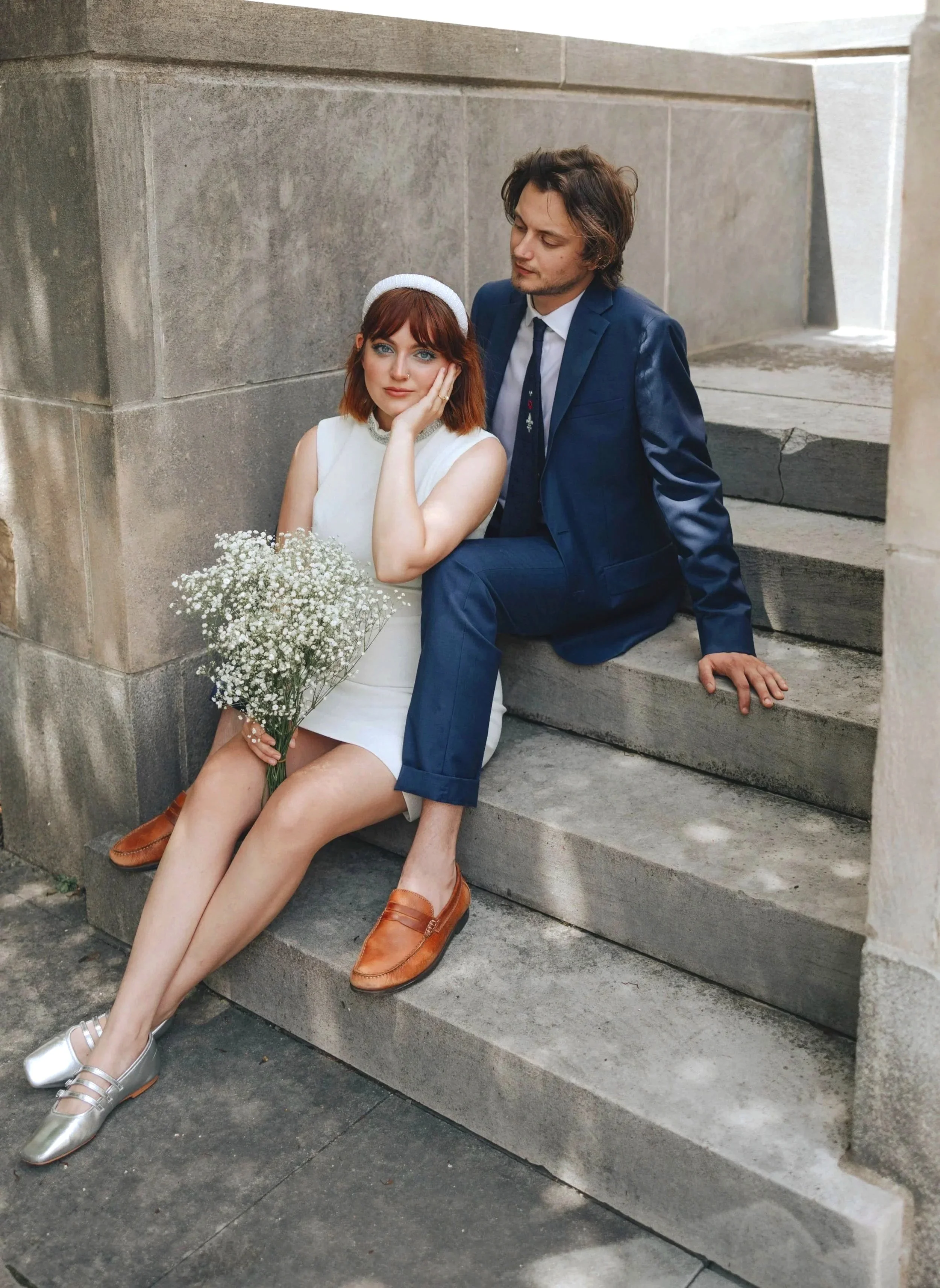 A woman in a white dress sitting on stairs holding a bouquet of flowers next to a man in a dark blue suit, both sitting on stone stairs. DOCUMENTARY STYLE, EDITORIAL STYLE