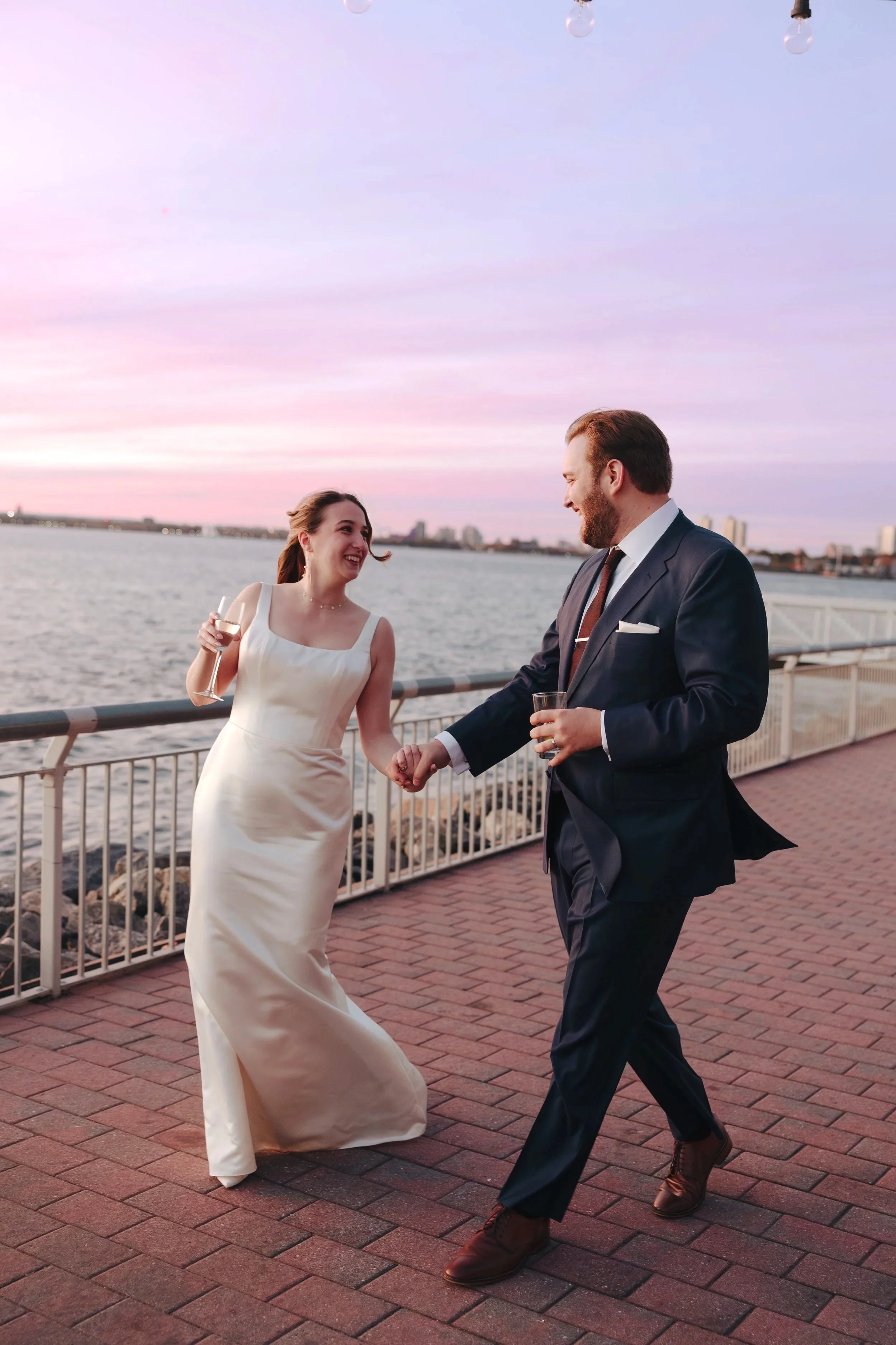 A man and woman holding hands and dancing on a waterfront promenade during sunset, both dressed in formal attire, with the woman wearing a cream-colored dress and the man in a dark suit, smiling at each other.