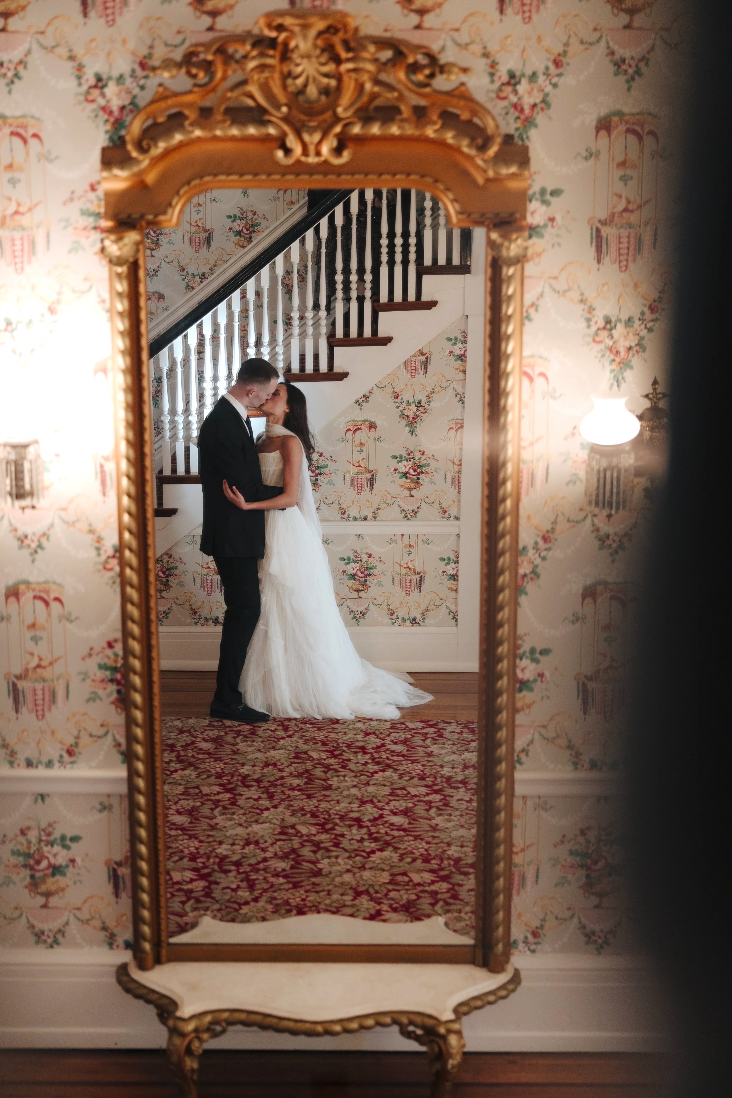 A couple dressed in wedding attire sharing a kiss in front of a staircase, viewed through an ornate antique mirror with floral wallpaper and a red rug in the background.