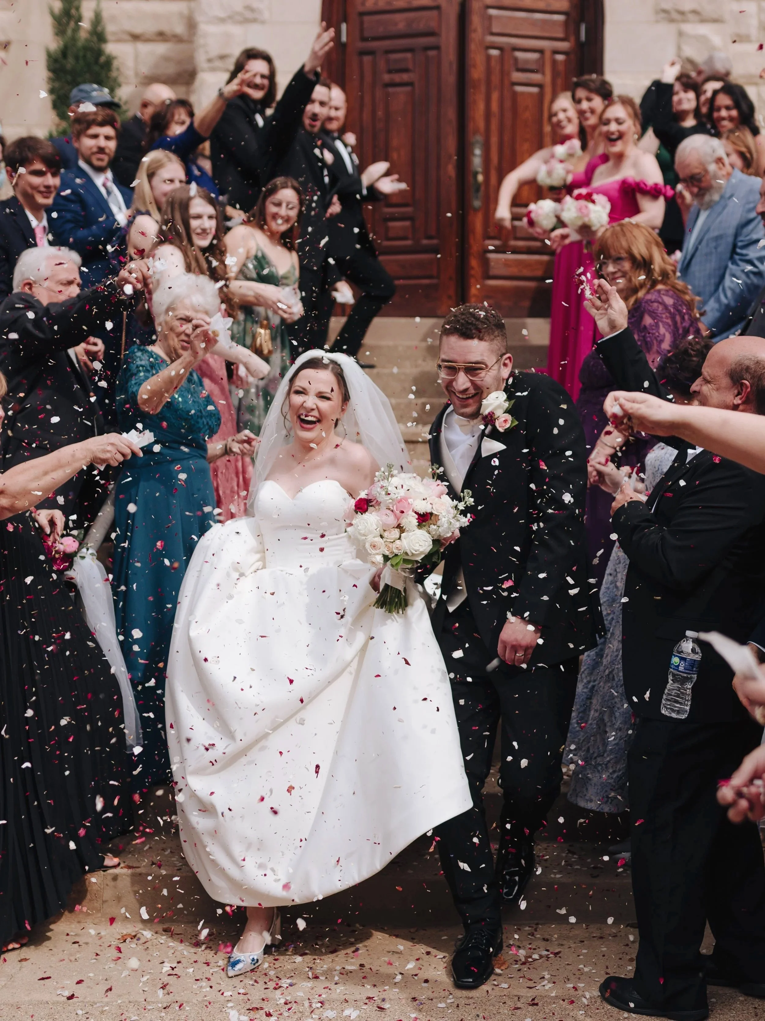 A newlywed couple is exiting a church surrounded by friends and family, celebrating with confetti and smiling. The bride is in a white wedding gown holding a bouquet, and the groom is in a black suit with a boutonniere.