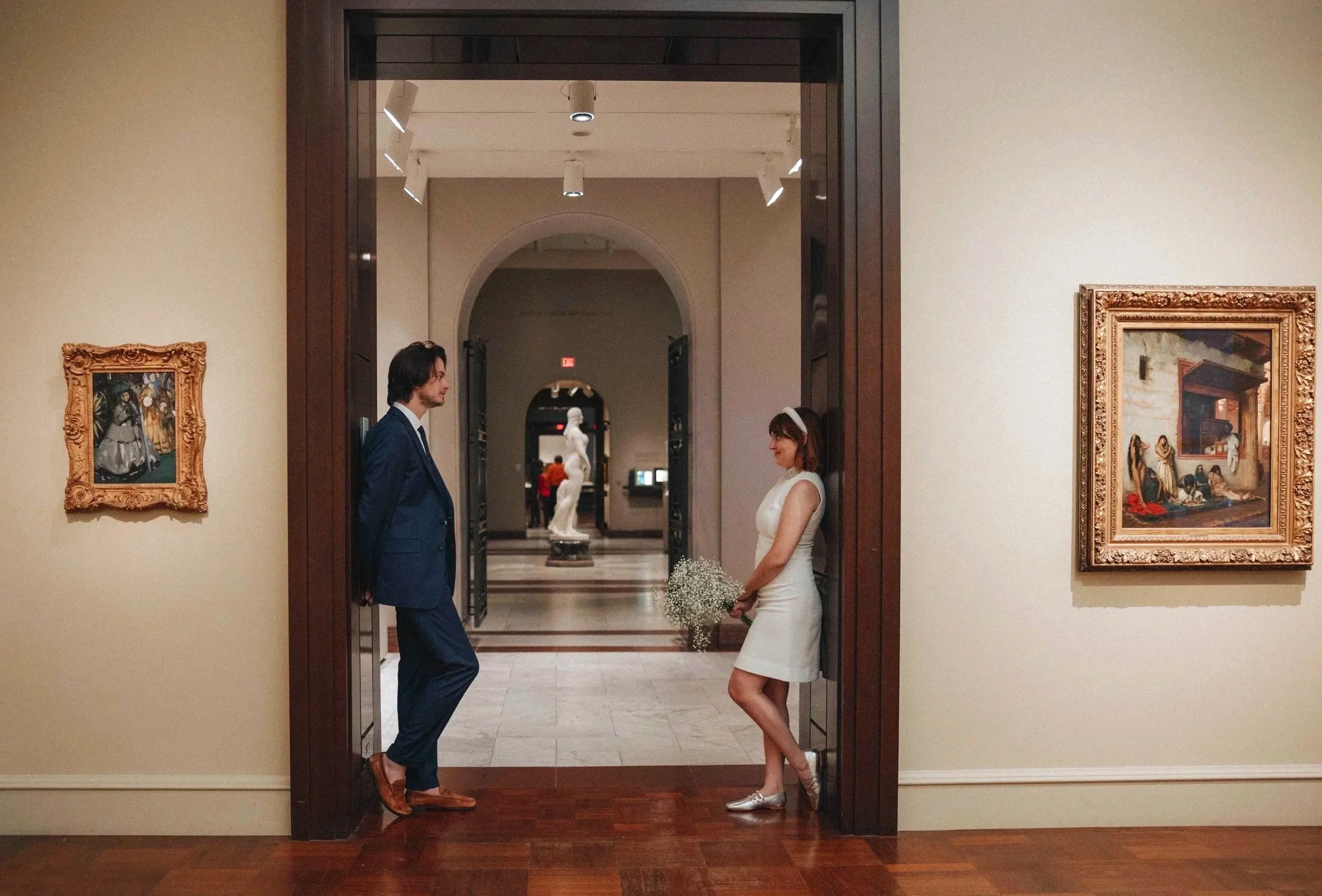 A man and a woman in a museum or art gallery talking to each other. The woman holds a bouquet of flowers and is dressed in a white dress, while the man is dressed in a blue suit. There are paintings on the walls and sculptures in the background. DOCU