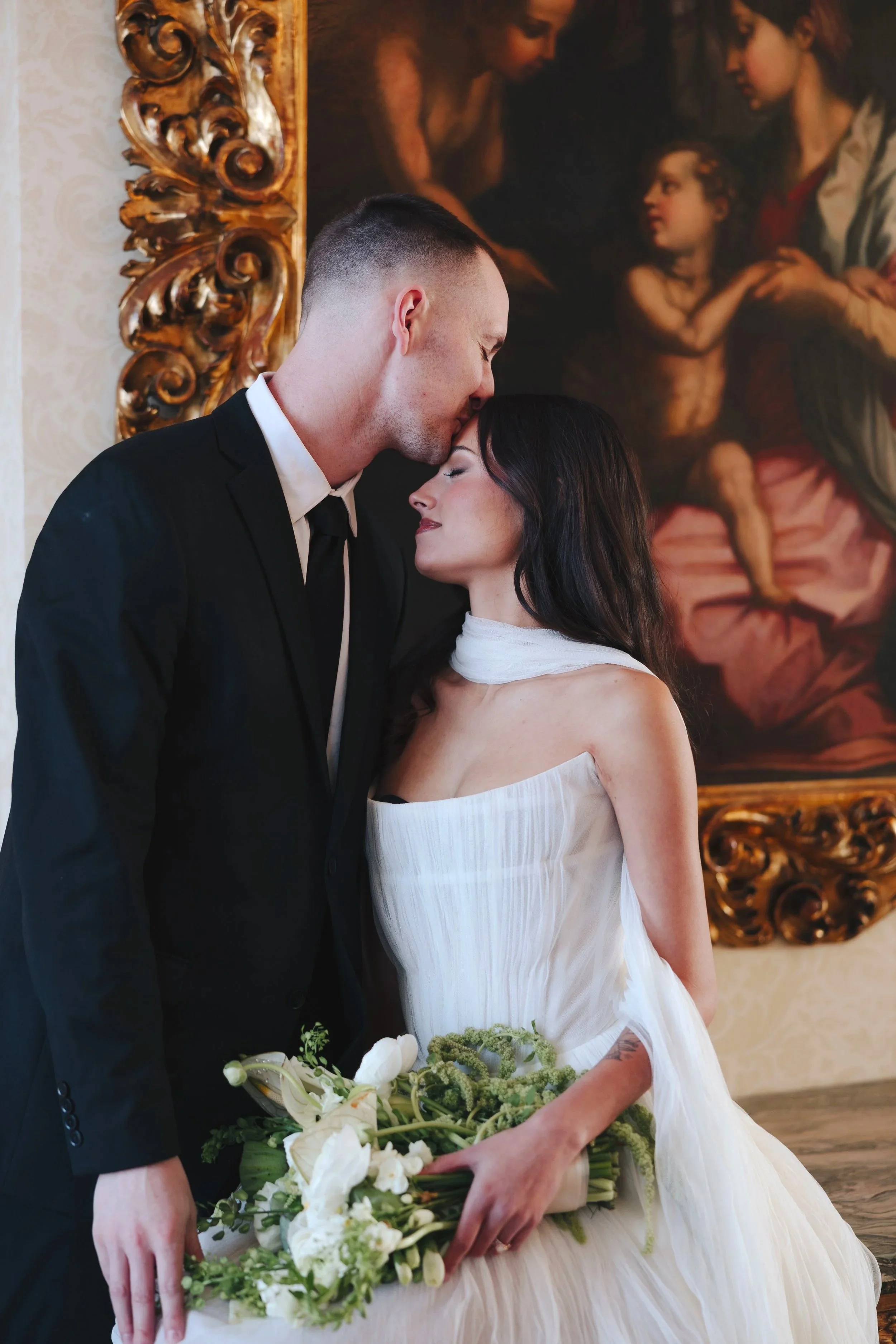 A newly married couple on their wedding day, with the groom kissing the bride on her forehead. The bride is holding a bouquet of white flowers and has long dark hair, while the groom is wearing a black suit and tie. They are indoors, with a large rel