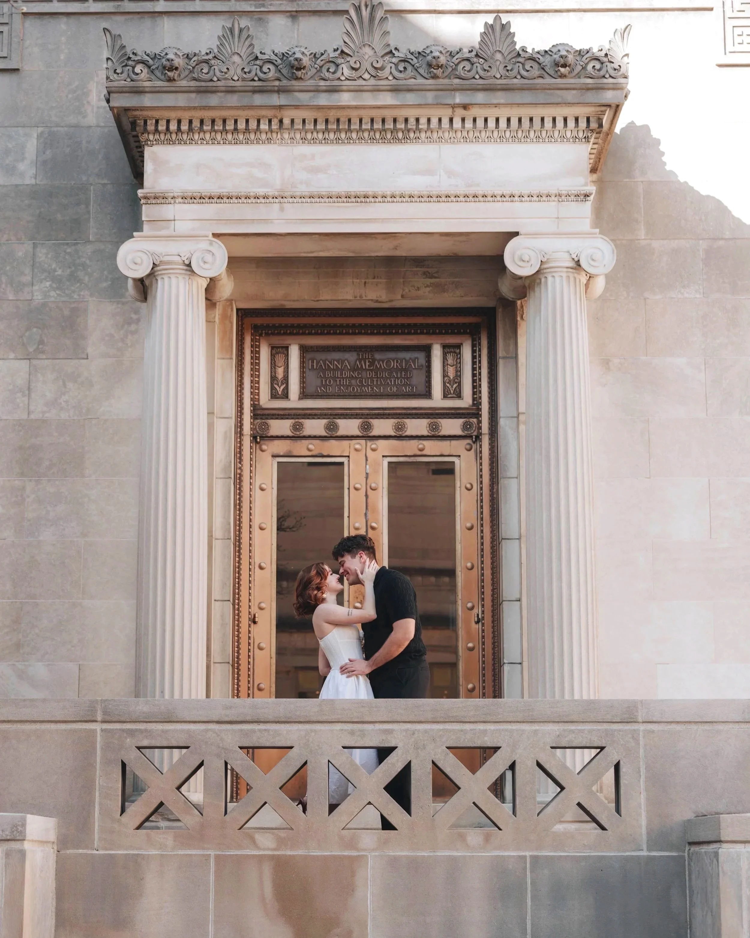 A couple standing close together in front of a historic building with ornate columns and decorative carvings, embracing and touching foreheads.