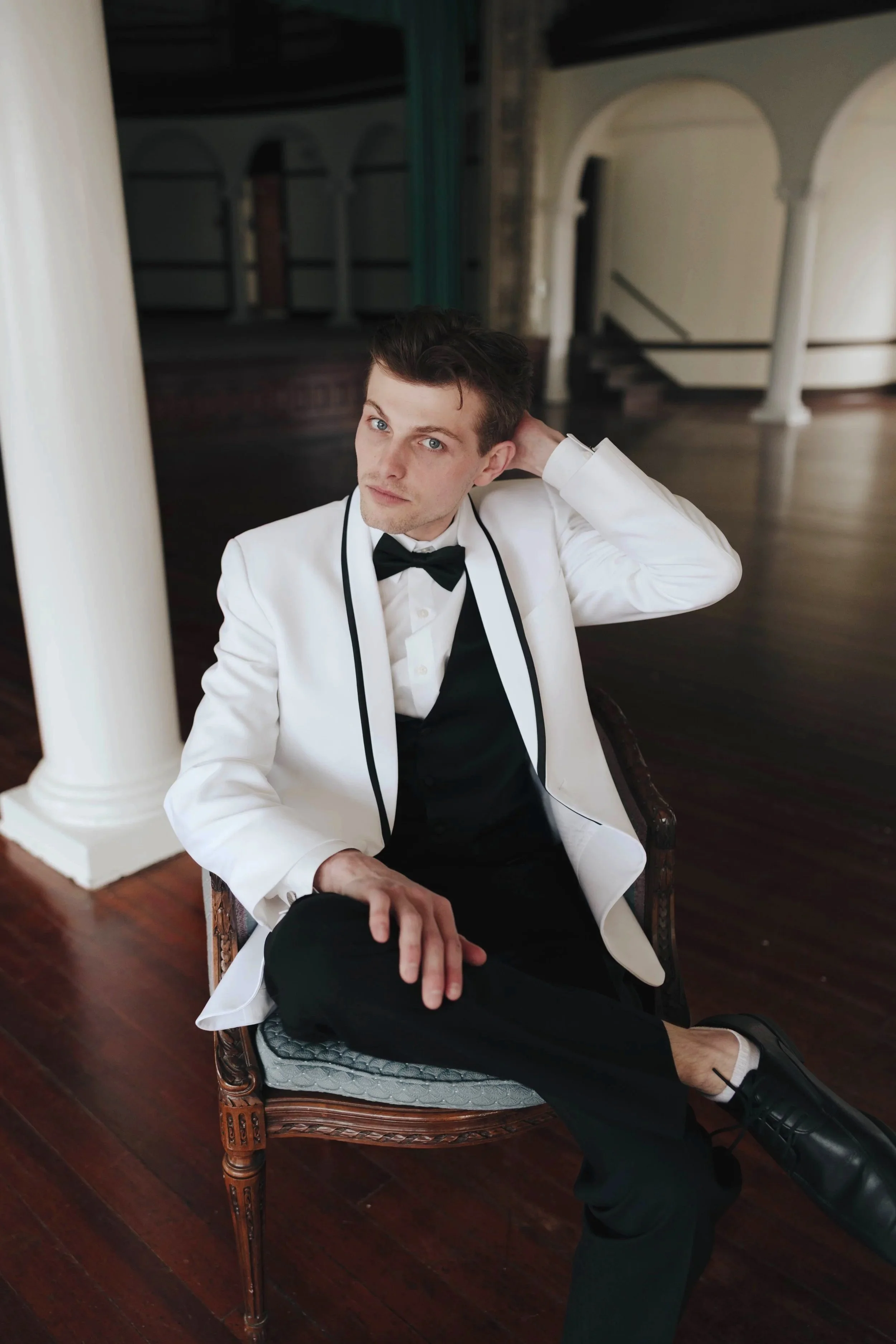 A young man in a white tuxedo jacket, black vest, and bow tie sitting on a vintage chair in an elegant, spacious room with white columns and wooden floors.