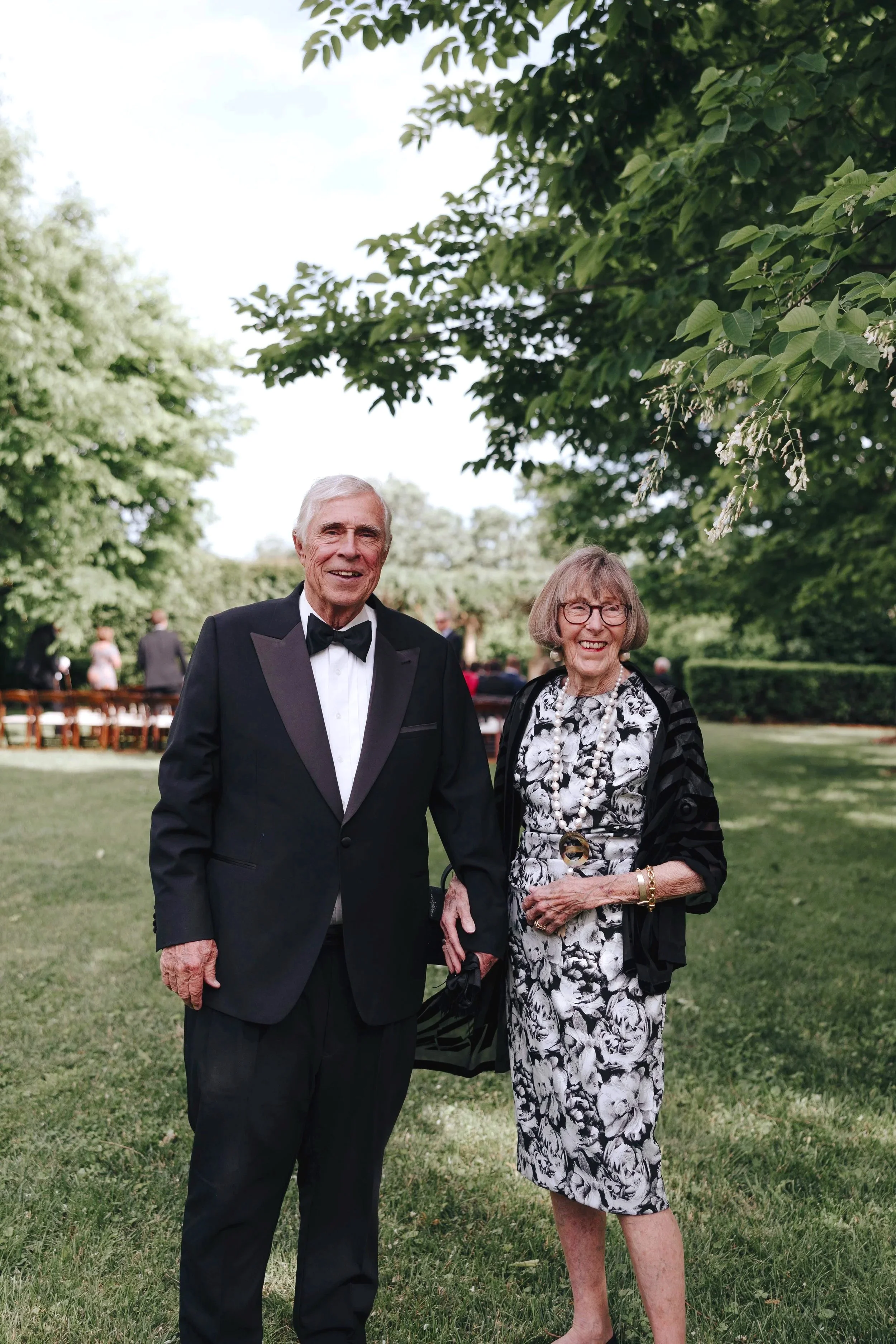An elderly man and woman dressed in formal attire standing on a grassy area outdoors, smiling for the camera with trees in the background.