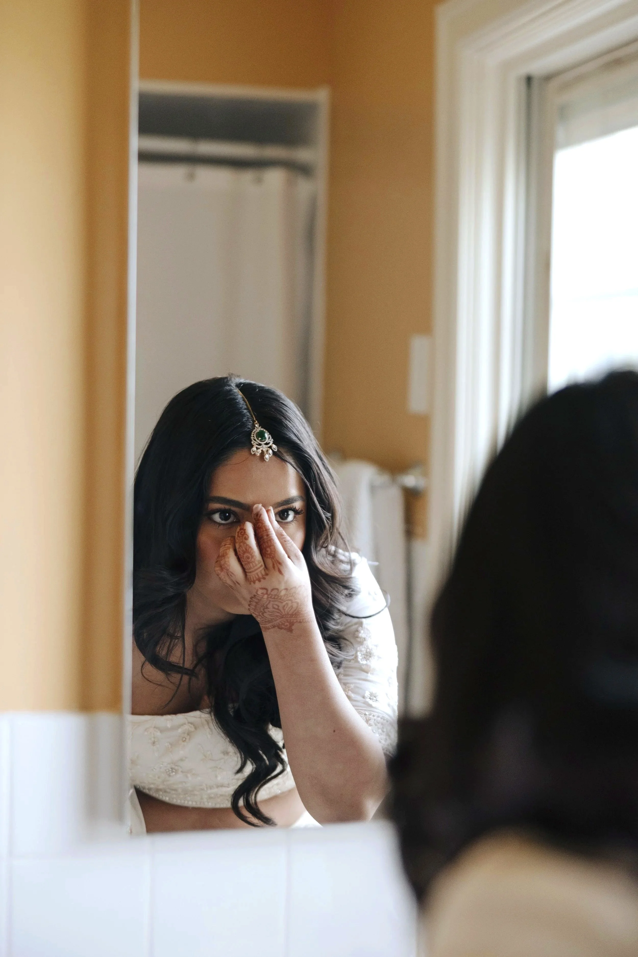 Young woman with dark hair applying makeup or adjusting appearance in front of a mirror, wearing traditional jewelry and clothing, with henna on her hand.