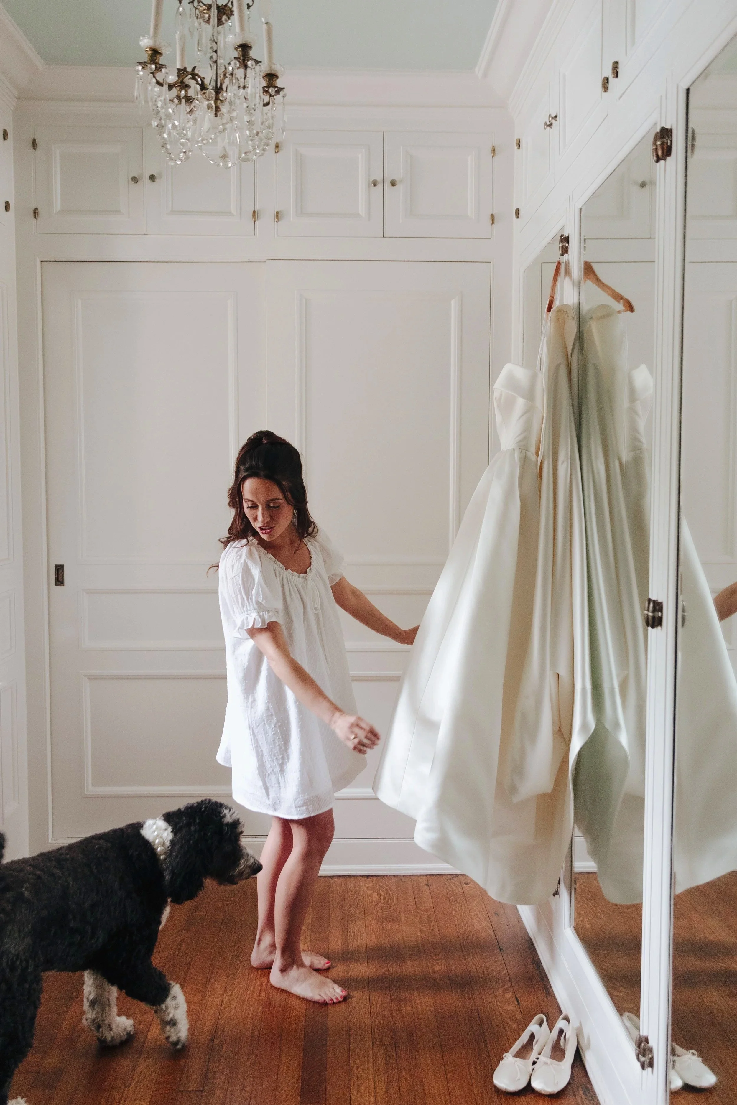 A woman standing in a room with white walls and a wooden floor, observing a wedding dress hanging on a mirror, while a black and white dog looks on.