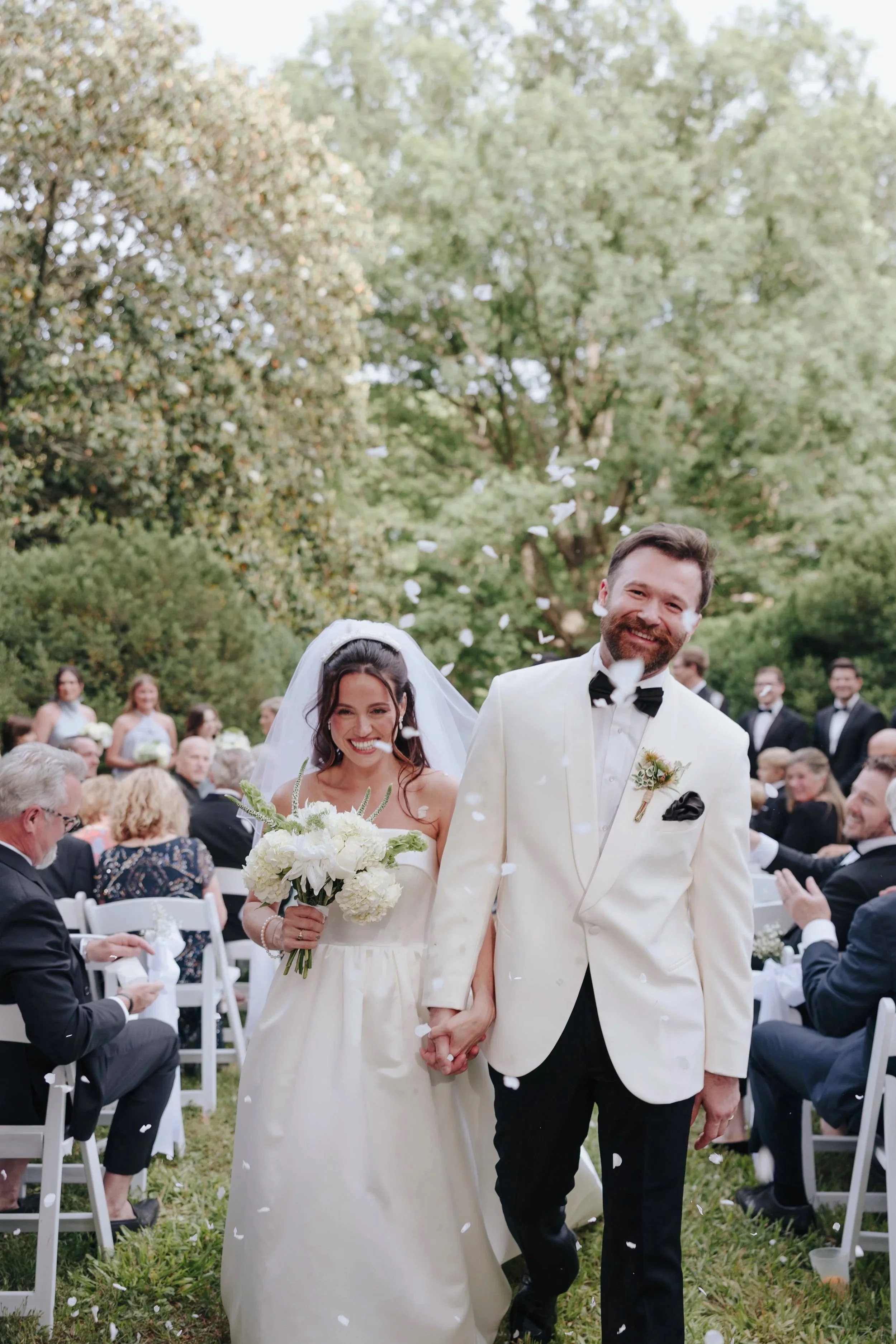 A bride and groom walking hand in hand at their outdoor wedding ceremony, smiling amid falling white flower petals, with guests seated in chairs and lush green trees in the background.