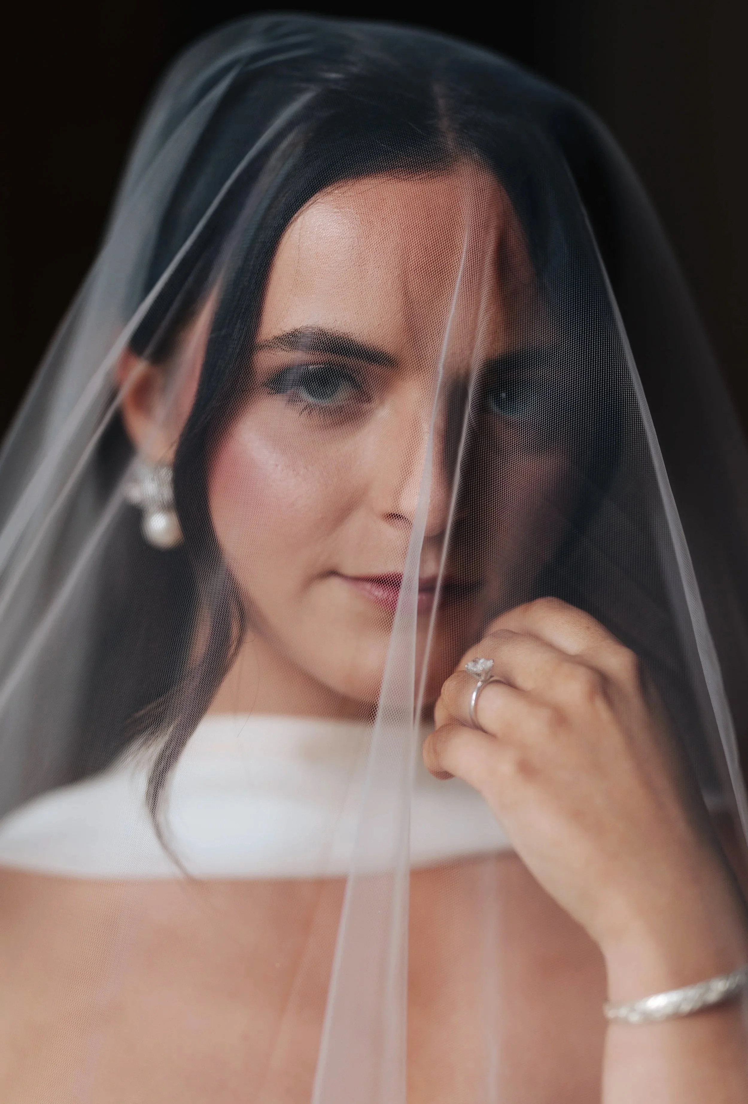 Close-up of a bride with dark hair and makeup, wearing a veil and a sparkling ring, smiling softly.