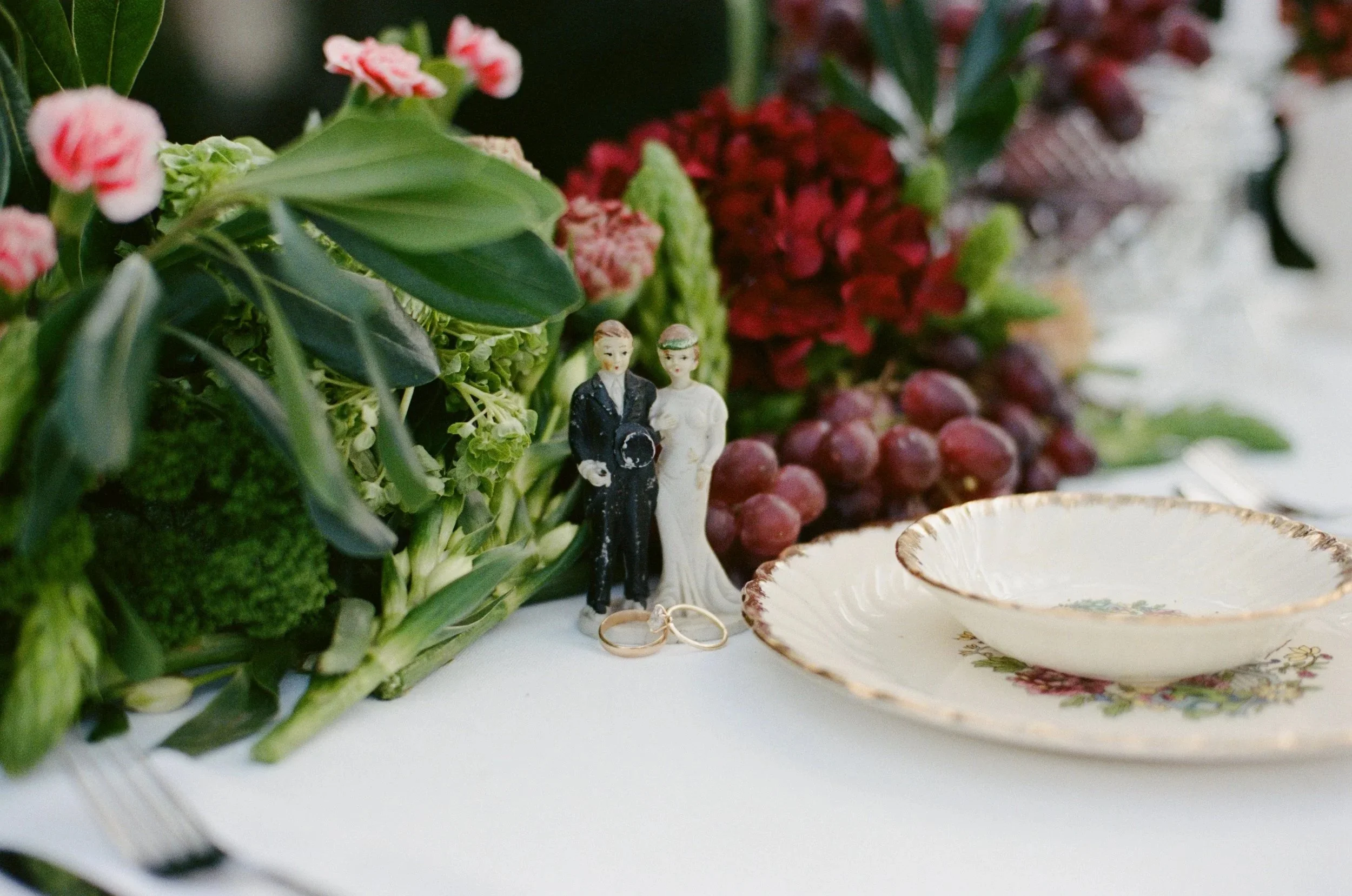 Miniature figurines of a man and woman standing among leaves and flowers, with a decorative plate and jewelry in the foreground.