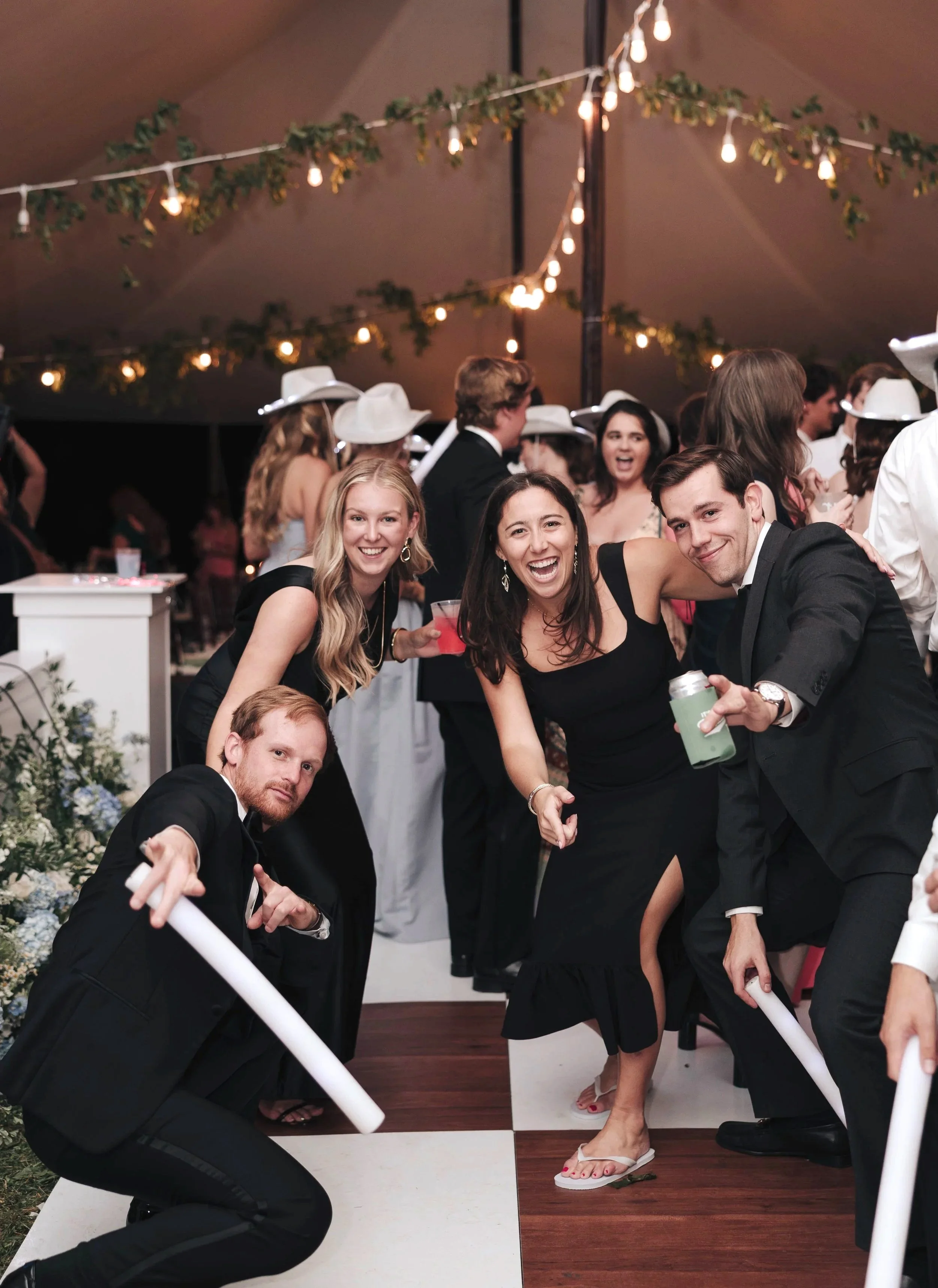 People celebrating at a party under a tent with string lights. Four individuals in the foreground are posing and smiling, holding drinks and props, with others dancing and chatting in the background.