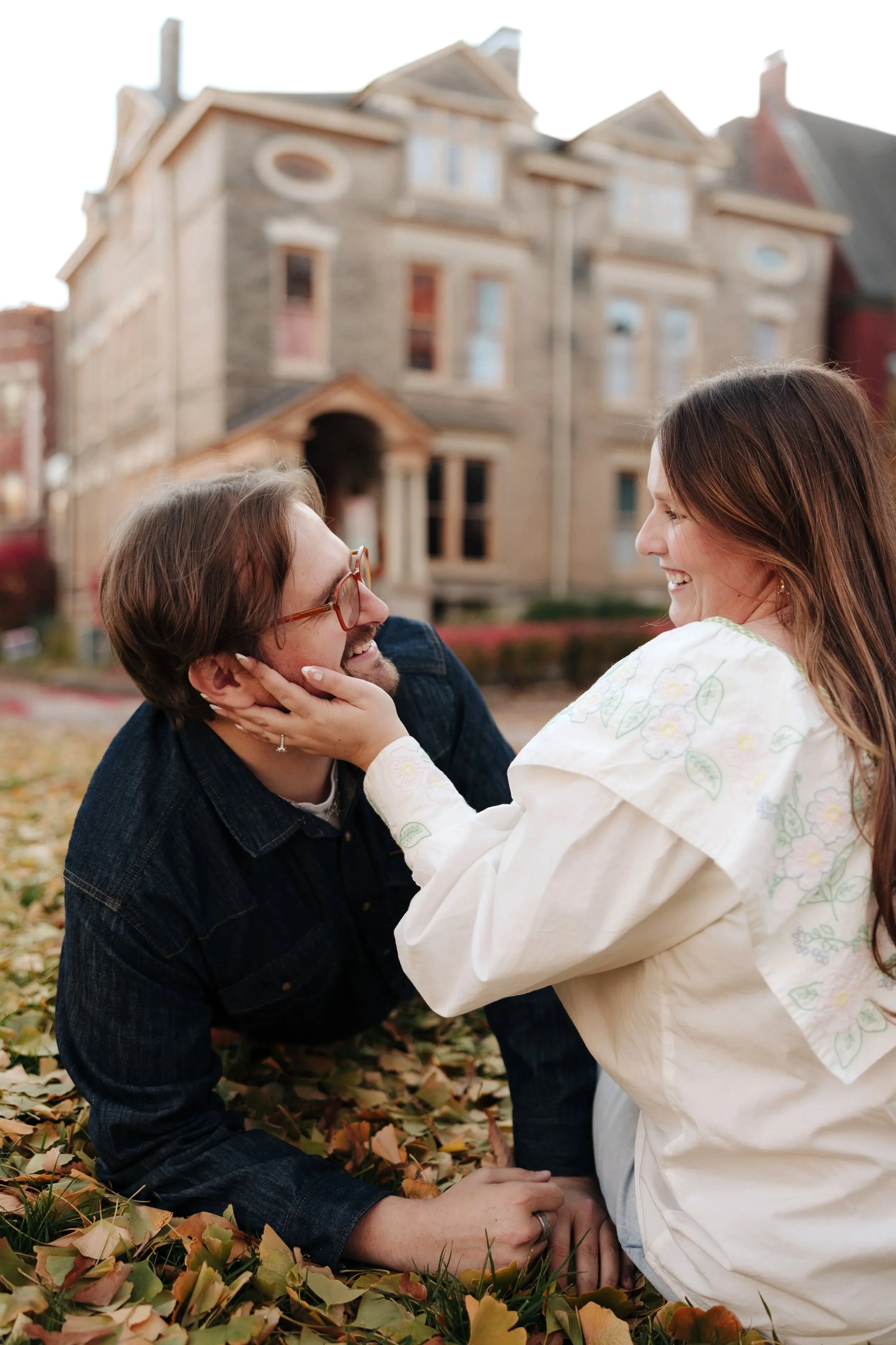 A couple playfully interacting outside on fallen autumn leaves with a large historic house in the background.