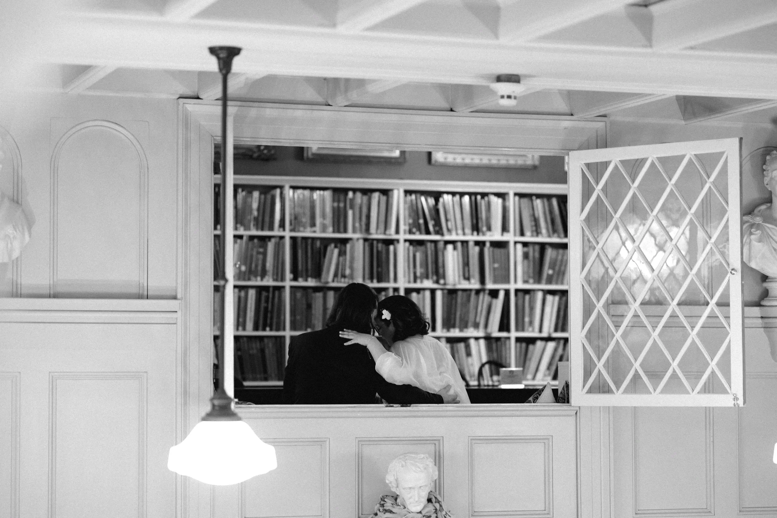 Two people sitting close together in a room with bookshelves, seen through a mirror with an open lattice window.