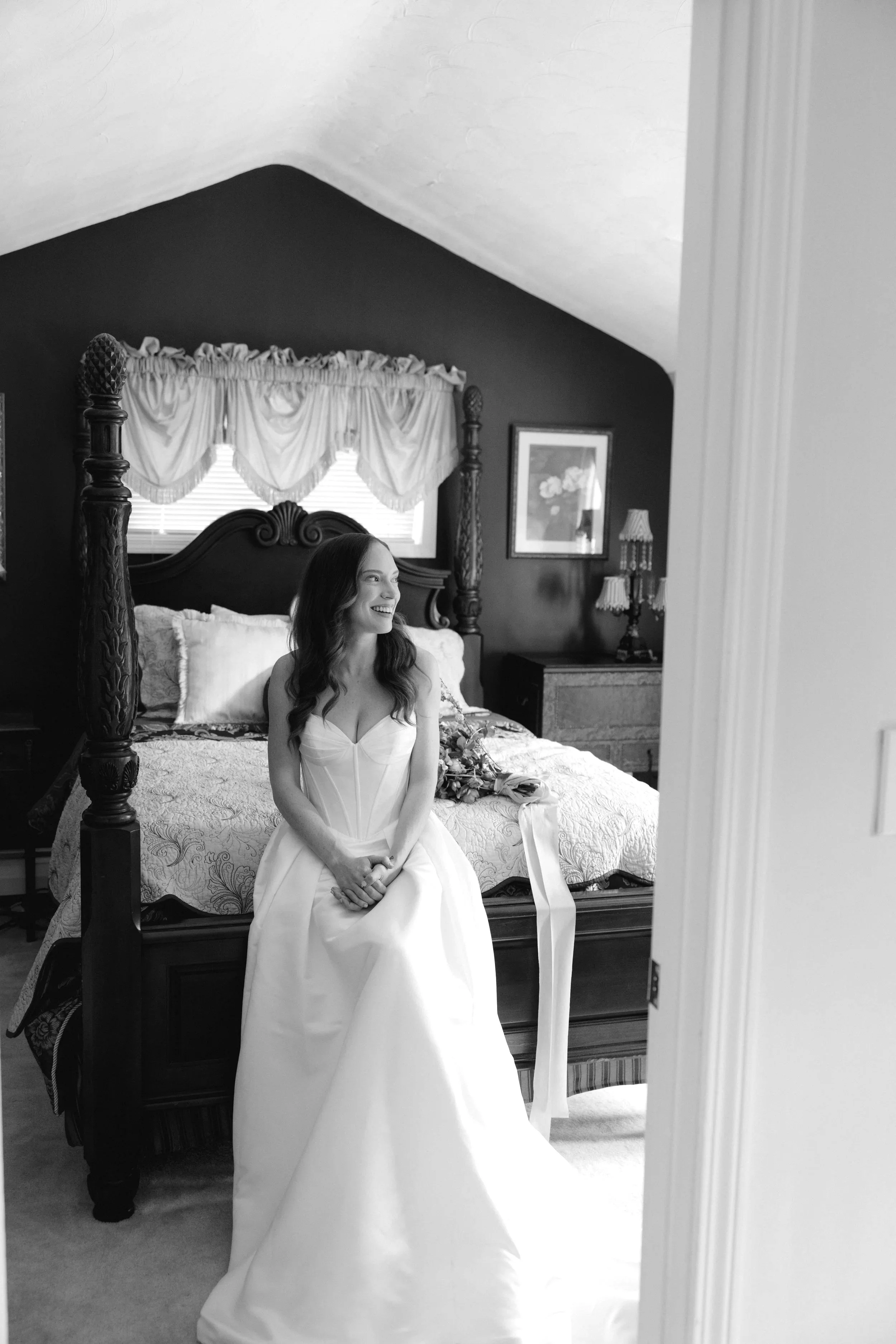 A woman in a wedding dress sitting on a bed inside a bedroom, smiling and looking to her right.