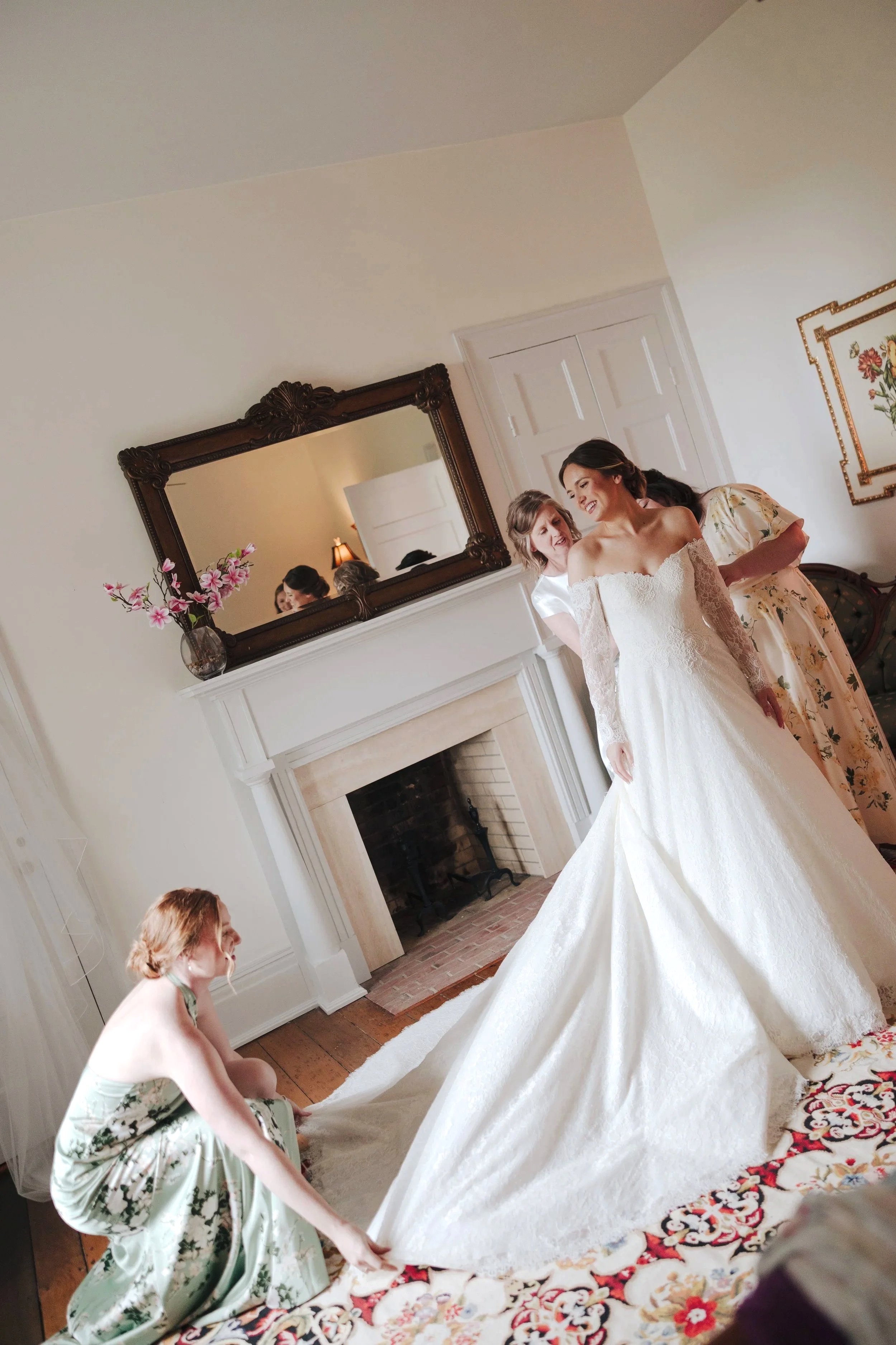 A bride in a white wedding dress is being assisted by several women, including one in a floral dress and another in a white satin blouse, as they prepare. The scene takes place in a living room with a fireplace, a large mirror above it, and a vase wi