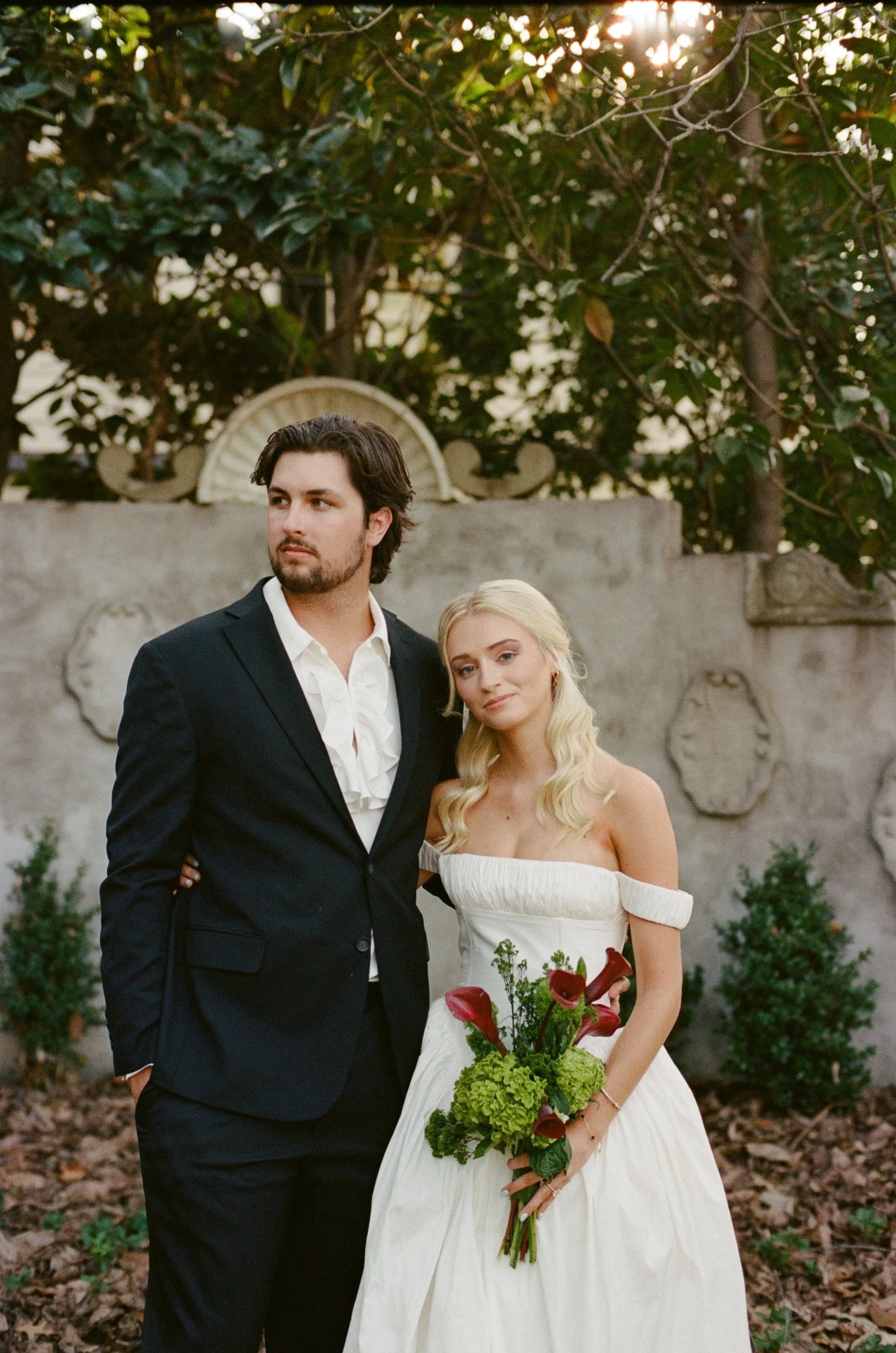 A couple dressed in wedding attire standing outdoors with a garden background, the man wearing a dark suit and the woman in a white wedding dress holding a bouquet of flowers.