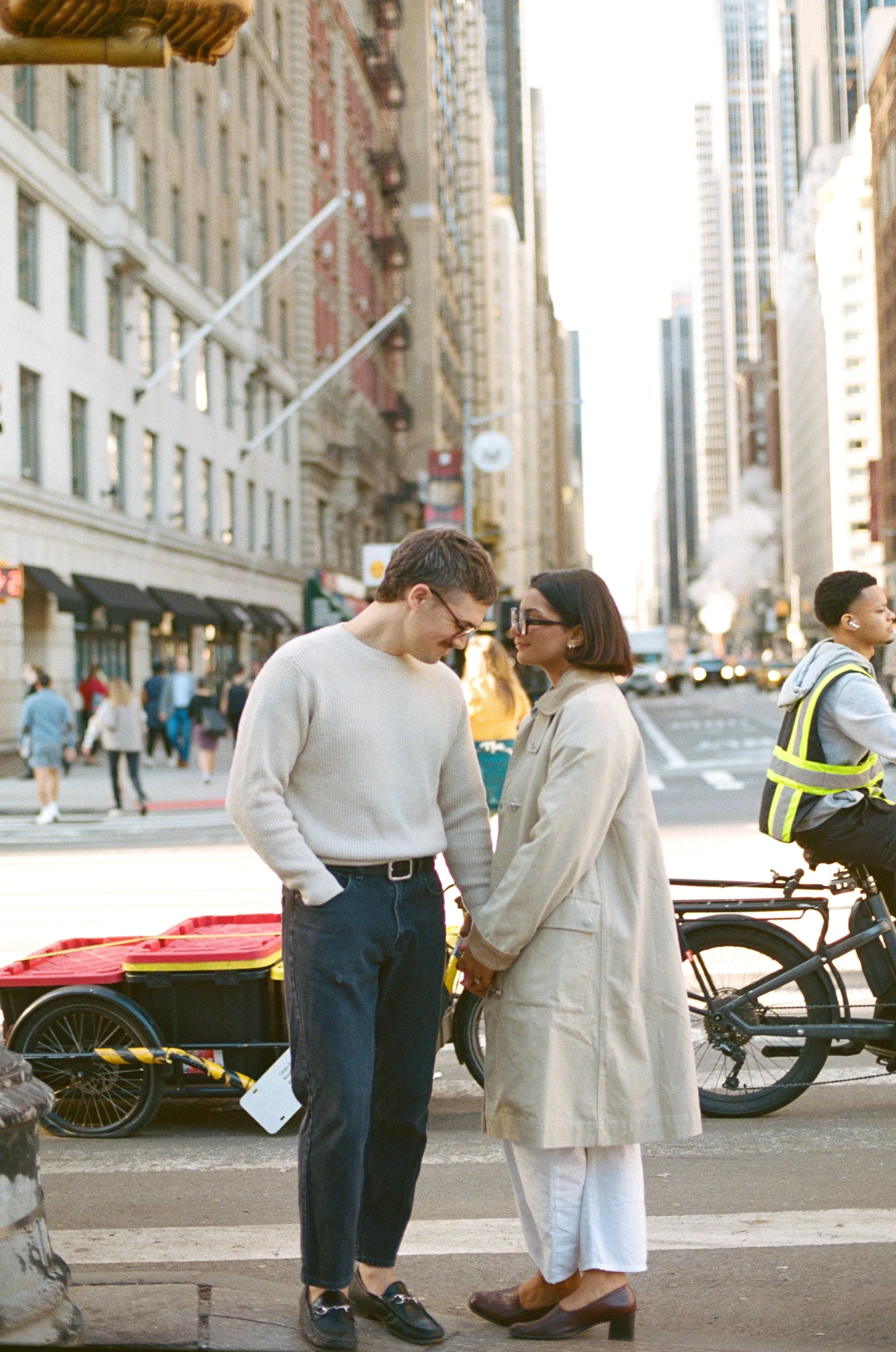A man and woman standing close together and looking at each other on a NYC city sidewalk, with tall buildings in the background. its a New York city photographer capturing them with a documentary candid approach