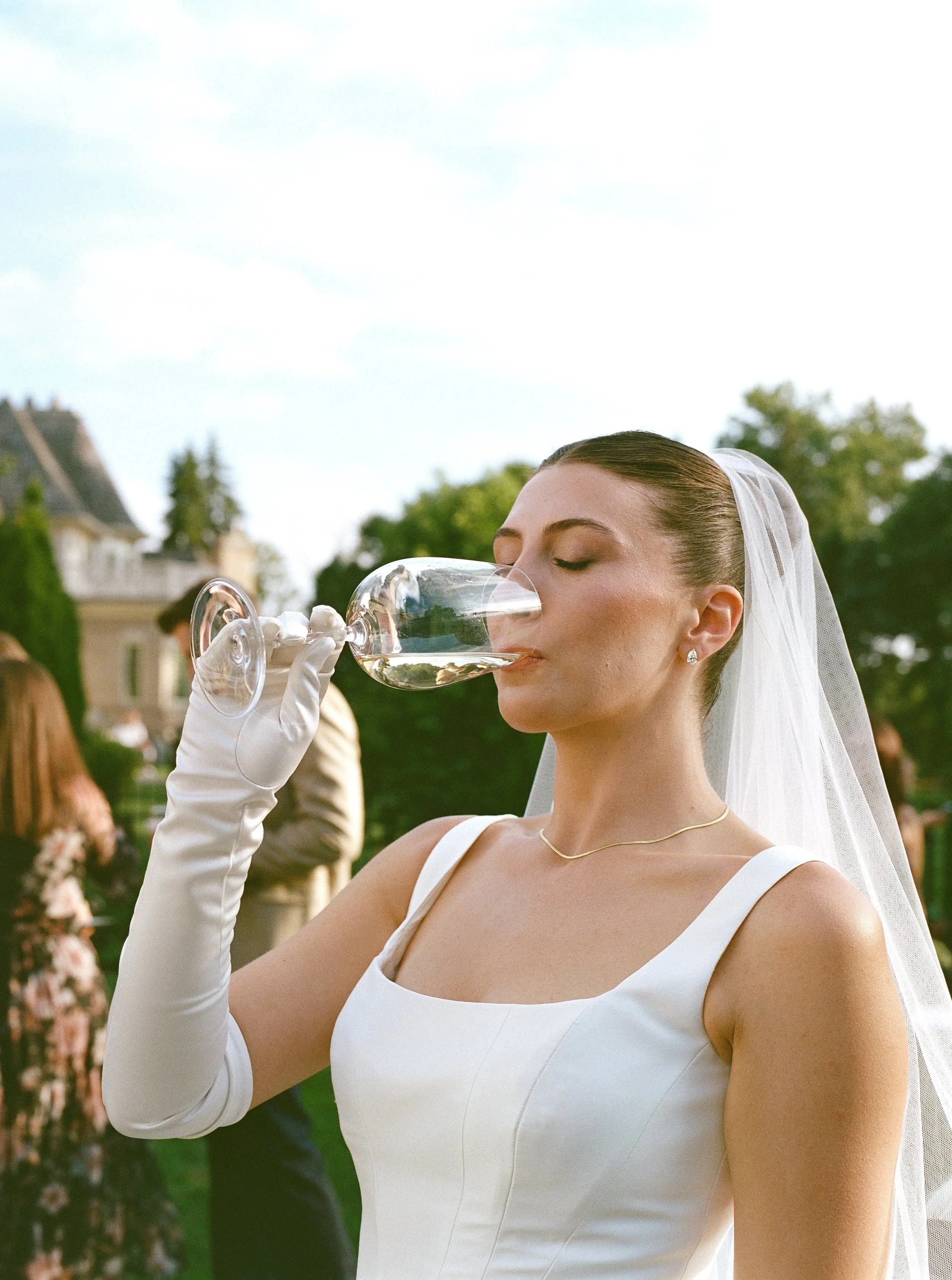 A bride in a white dress with a veil and gold necklace is drinking from a wine glass at an outdoor wedding reception.