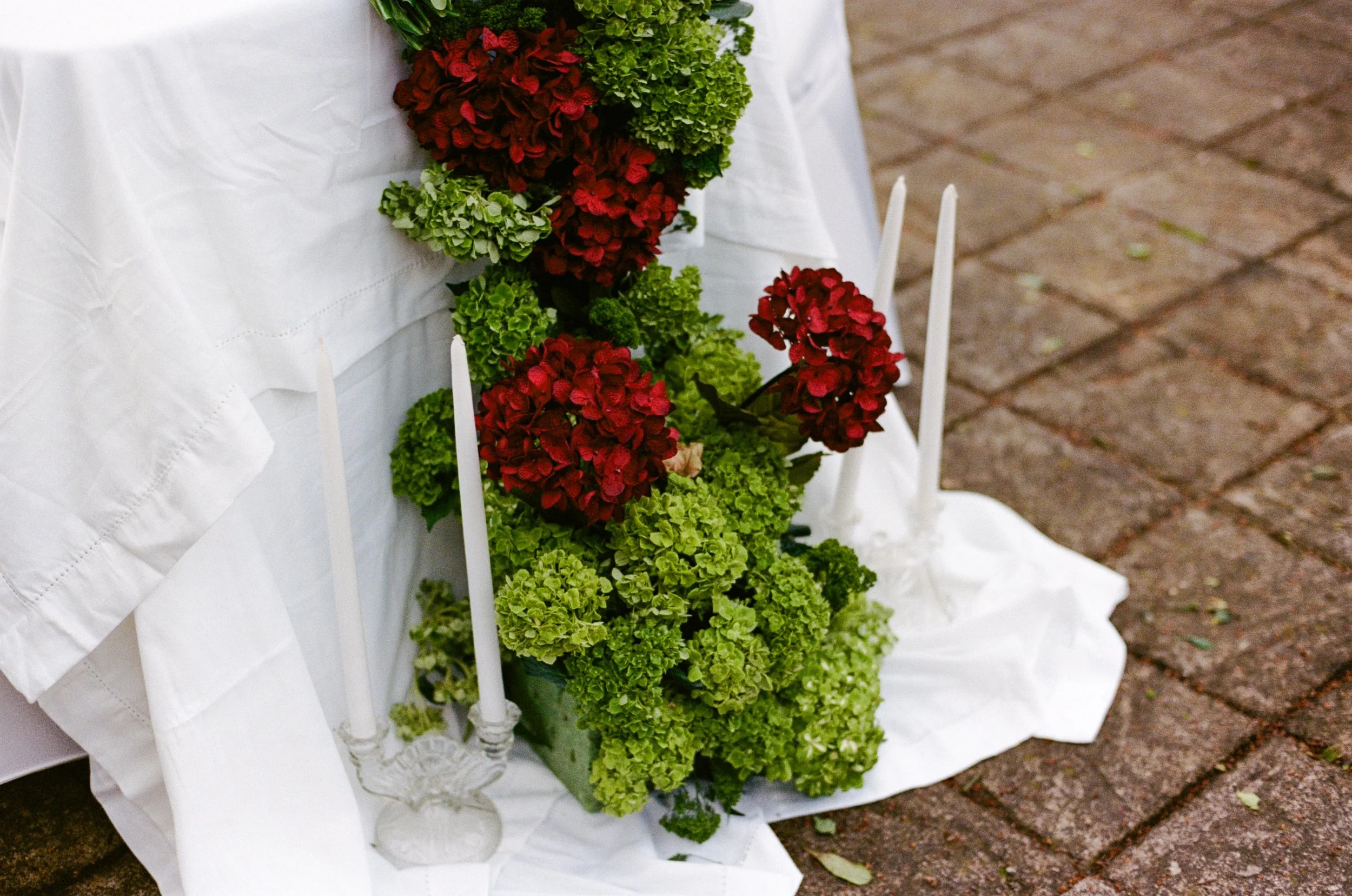 Arrangement of green and red hydrangeas with white candles on a white cloth on a brick ground.