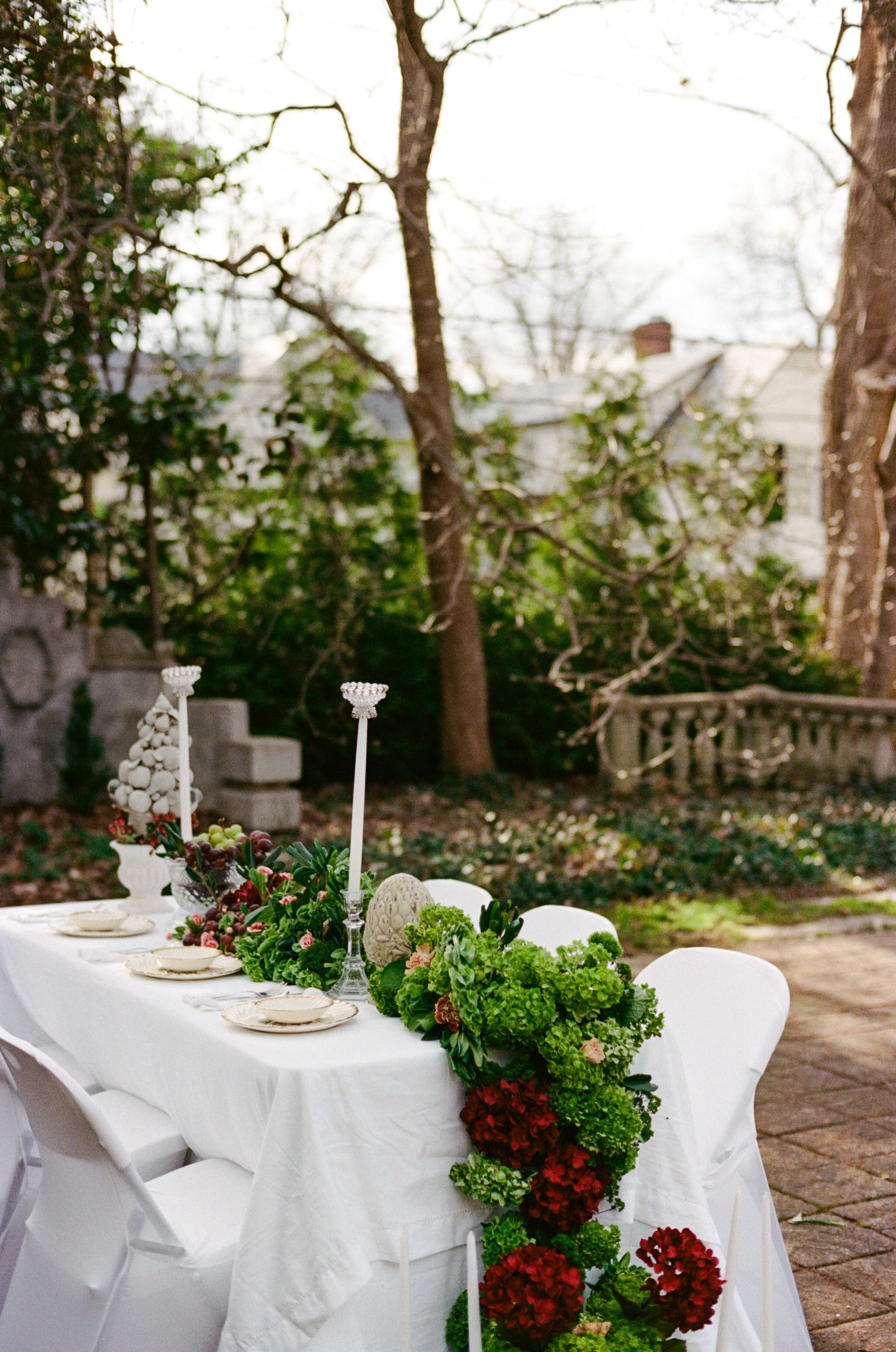 A decorated outdoor dining table with white tablecloth, surrounded by white chairs, featuring floral arrangements and tall white candlesticks, set in a garden with trees and a stone wall.