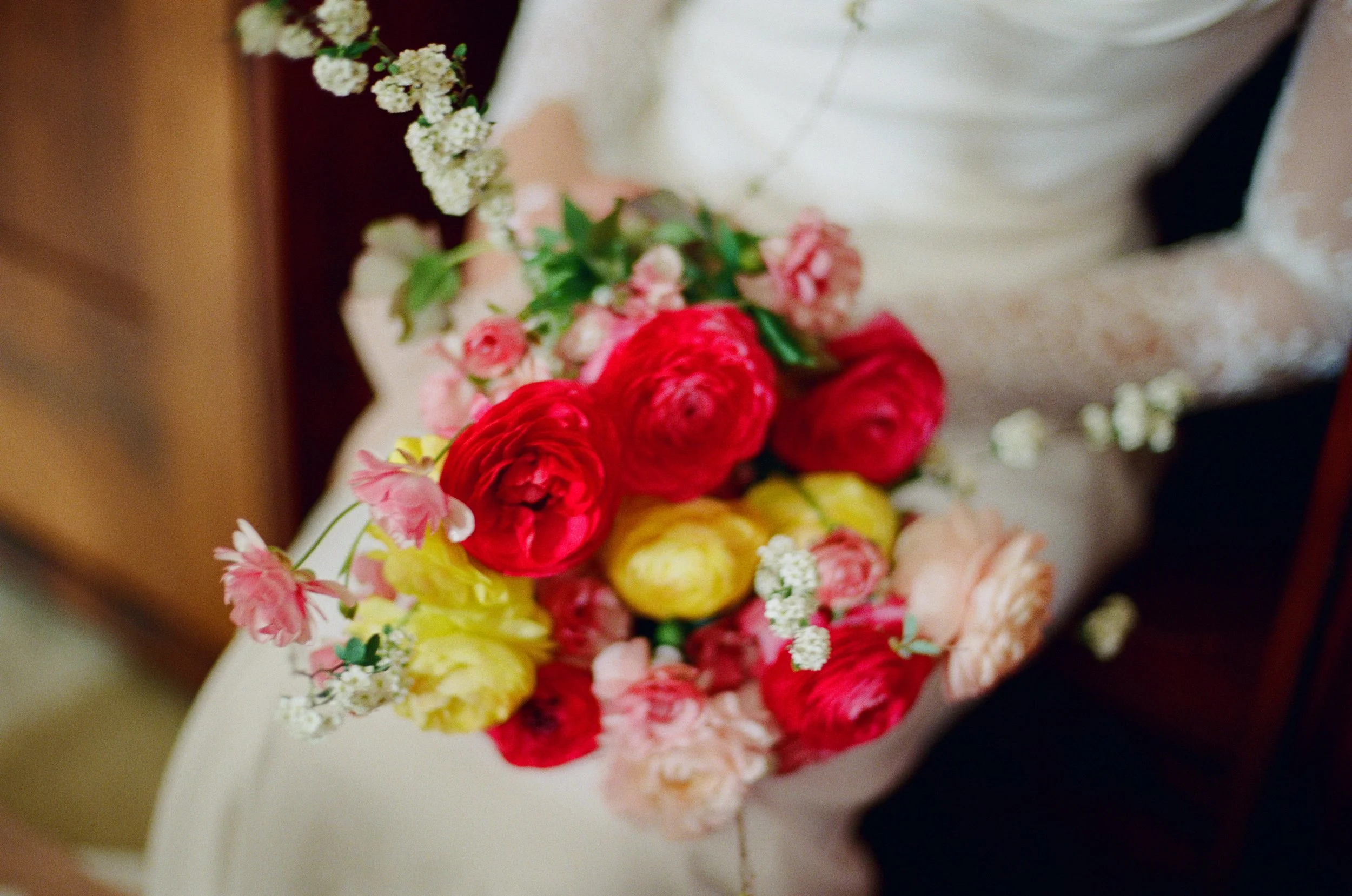 Close-up view of a bouquet of colorful flowers including red roses, pink carnations, and small white blossoms, arranged on a white surface with a blurred background.