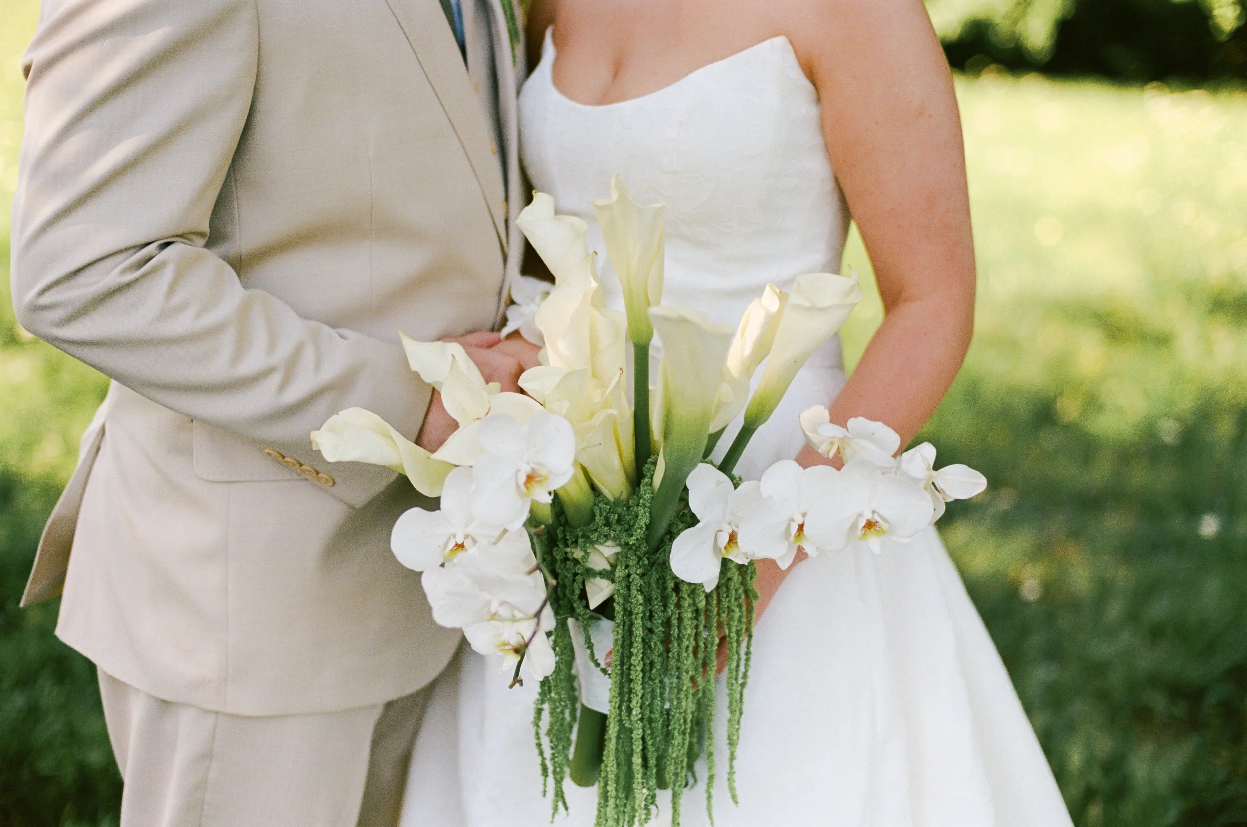 Close-up of a bride in a white dress holding a bouquet of white and green flowers, next to a groom in a beige suit. The background is blurred greenery.