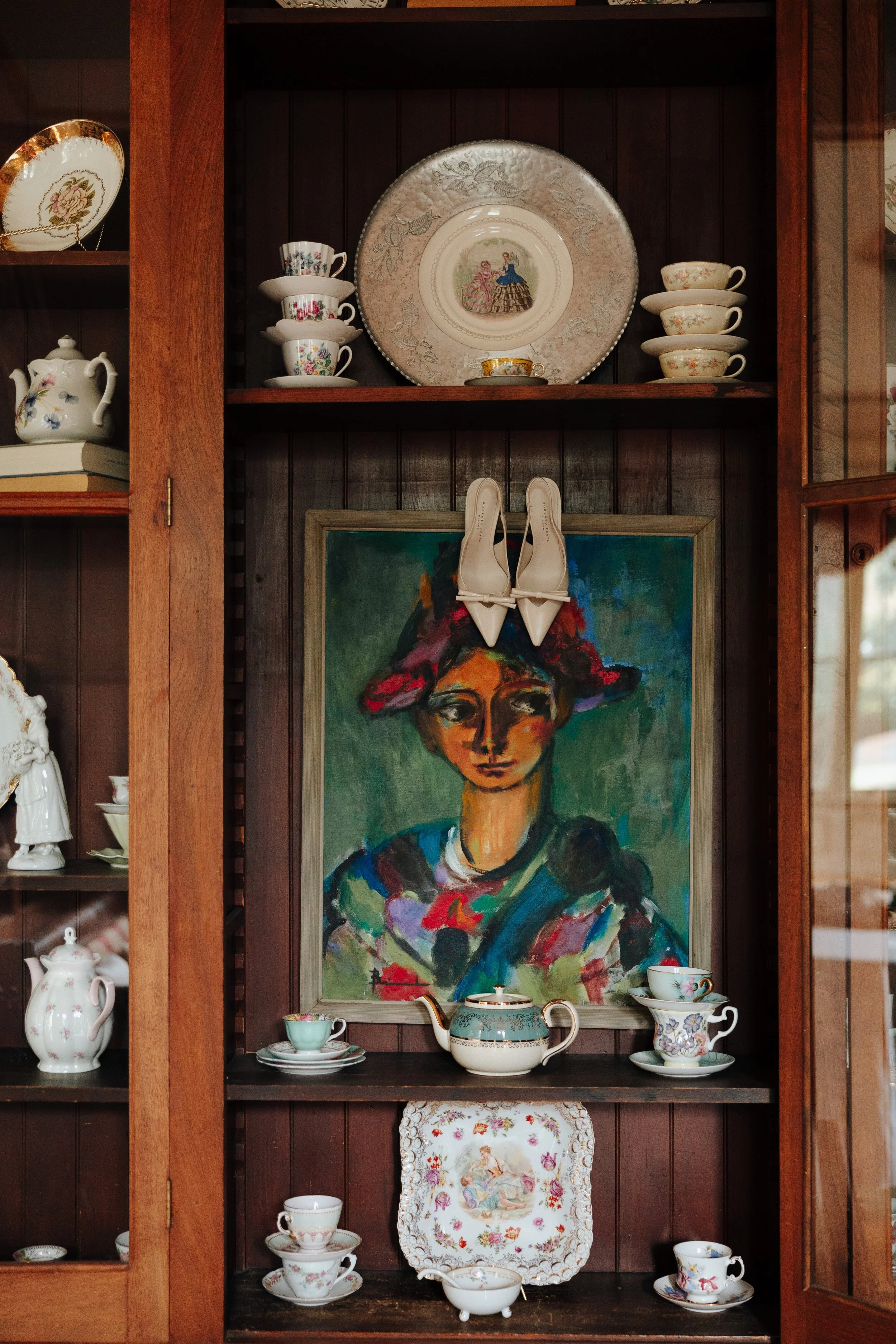 A wooden display cabinet containing fine china, a decorative plate with a Victorian scene, and a colorful portrait of a woman with a large hat.