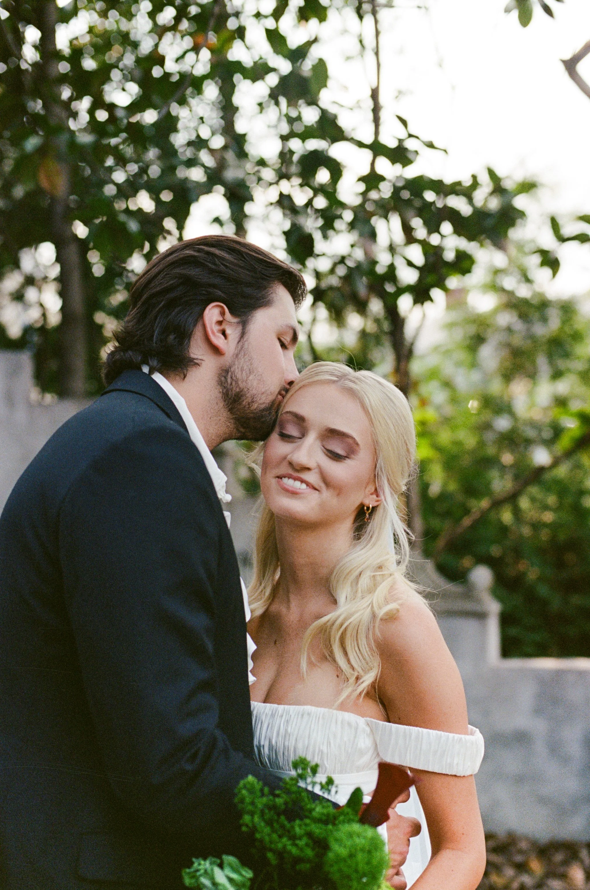 A romantic moment between a bride and groom outdoors, with the groom kissing the bride on her forehead, and the bride smiling with her eyes closed, surrounded by greenery.