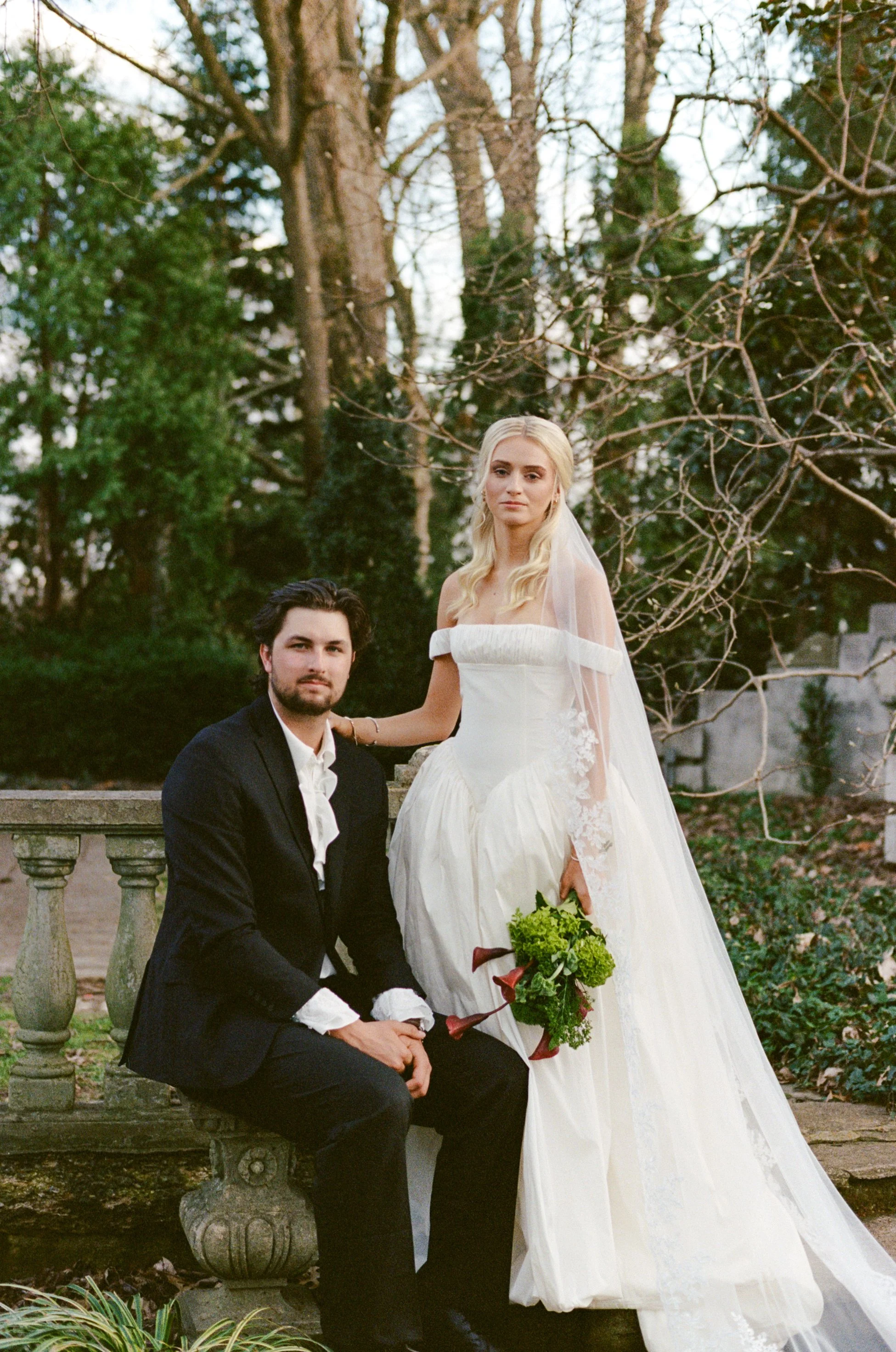 A bride in a white wedding dress holding a bouquet, and a groom in a black tuxedo with a white shirt, posing outdoors on a stone bench in a garden with trees and greenery.
