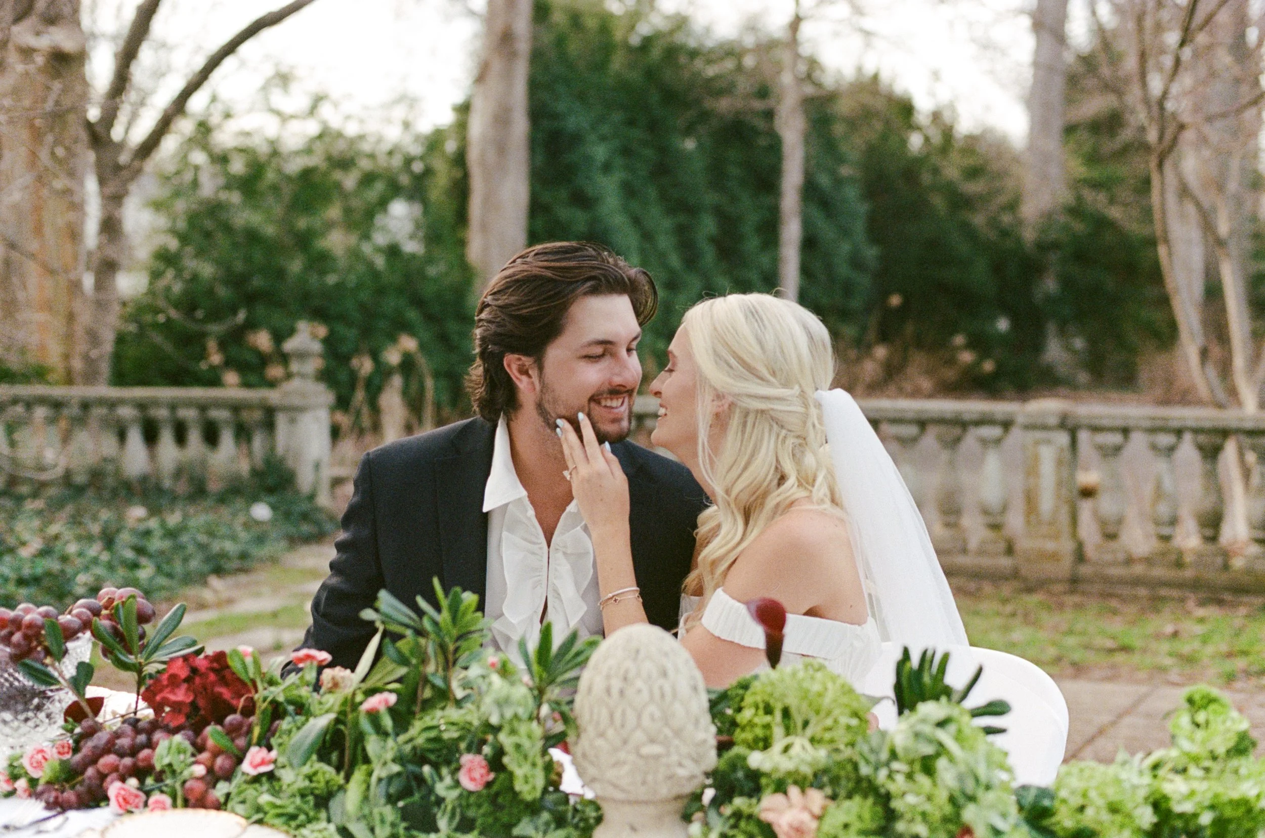 A newlywed couple shares a moment at their outdoor wedding reception, surrounded by greenery and decorated with flowers and a carved stone centerpiece.