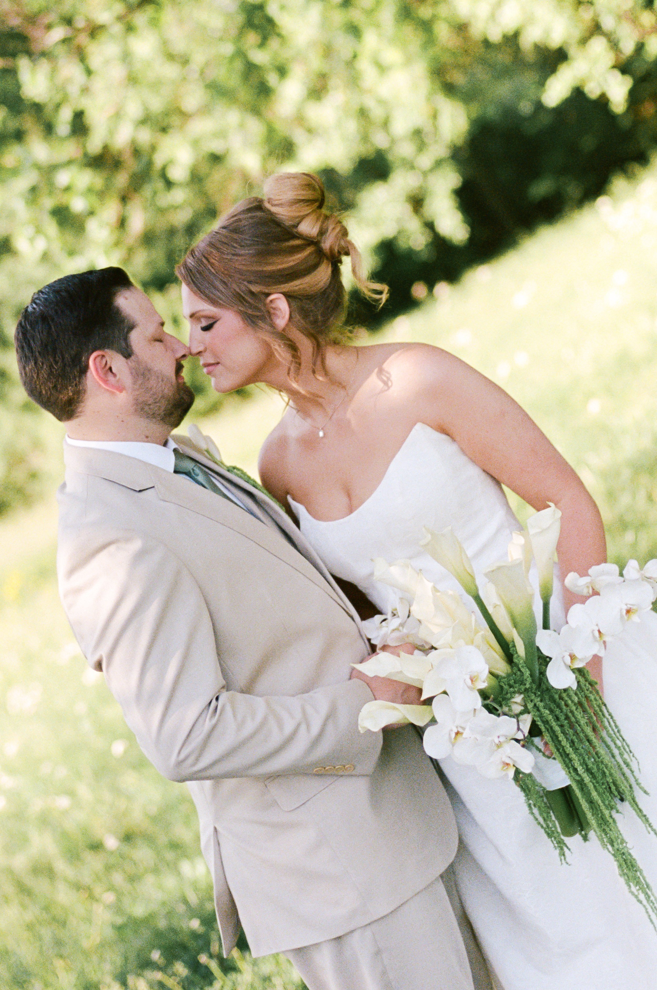 A bride and groom embrace outdoors, with the groom holding a bouquet of white orchids and greenery, against a background of green trees and sunlight.