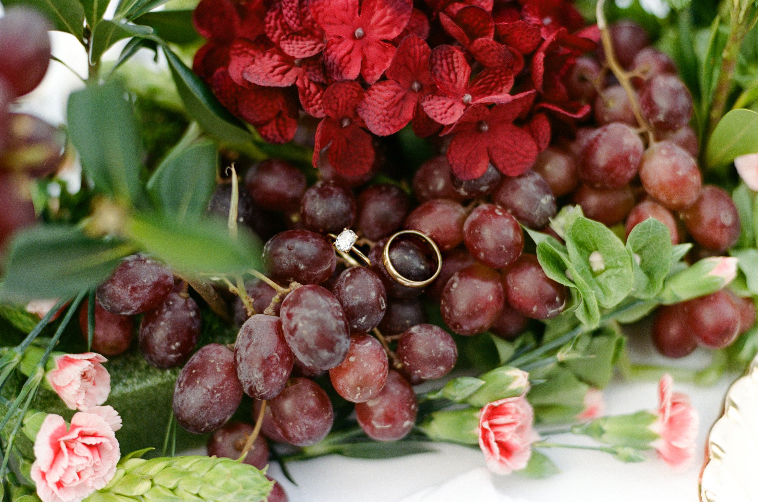 Close-up of a floral arrangement with red hydrangea, grapes, pink carnations, and greenery, with a gold ring and diamond ring resting on the grapes.