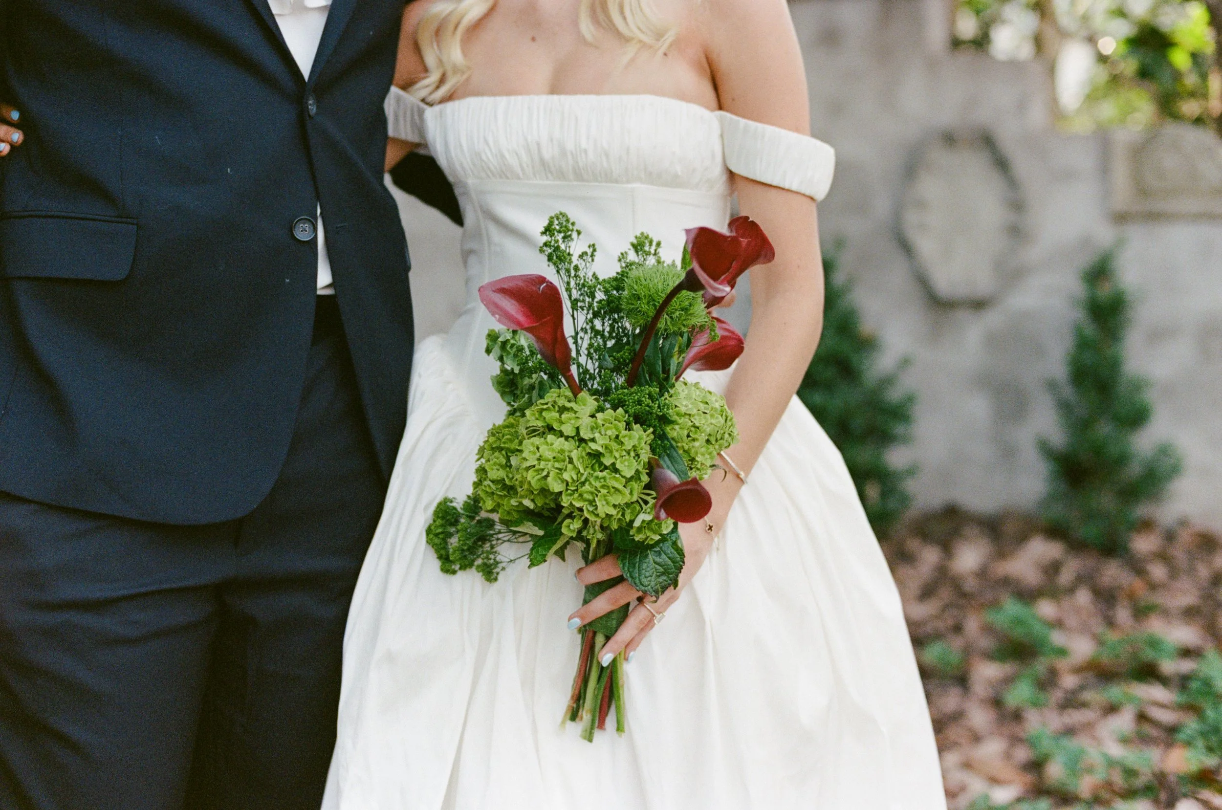 Close-up of a bride in a white wedding dress holding a bouquet of green and red flowers, standing next to a groom in a dark suit, outdoors with trees and a stone wall in the background.