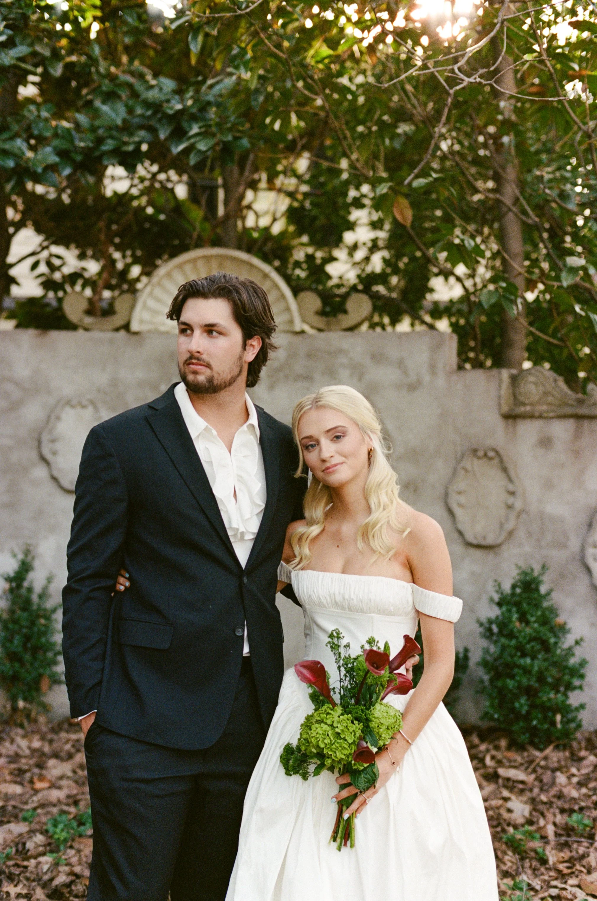 A young couple dressed in wedding attire standing outdoors in front of a stone wall with decorative plaques, surrounded by trees and greenery.