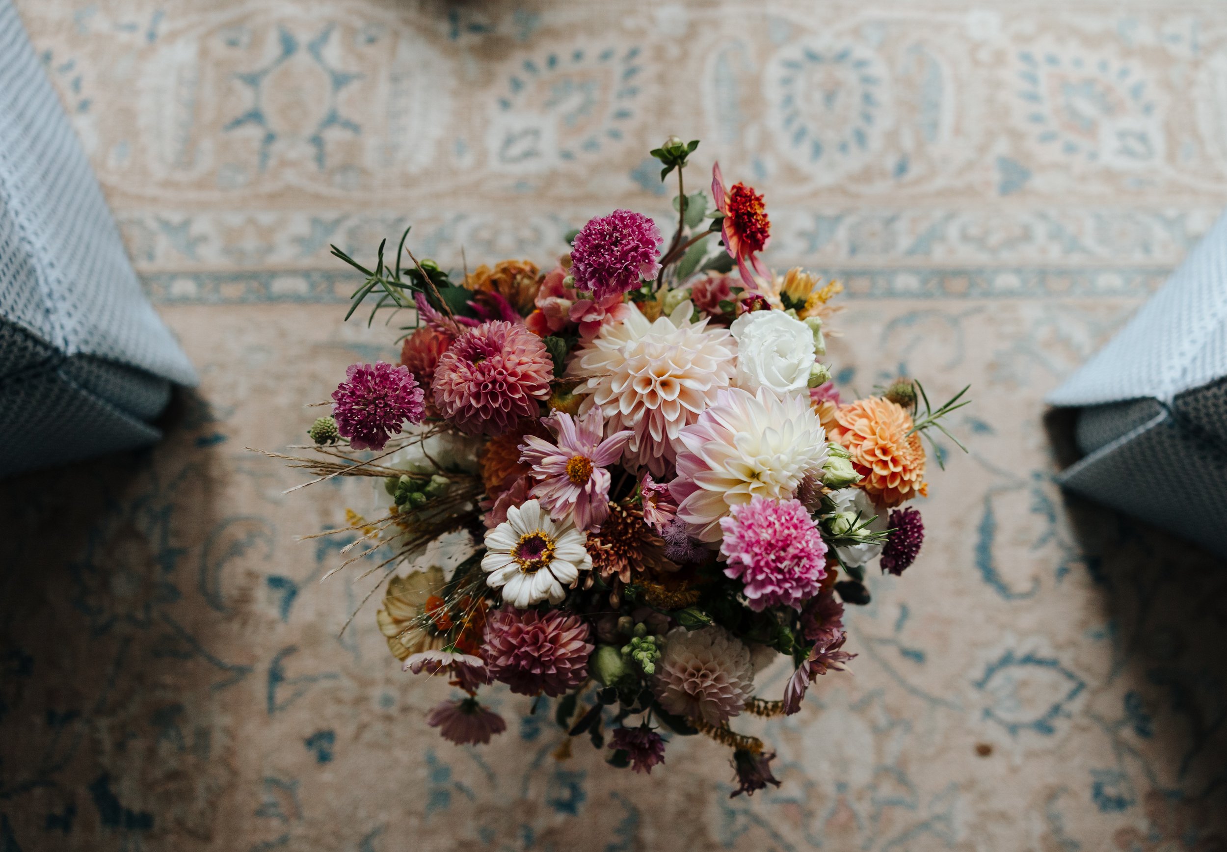 A colorful bouquet of various flowers including dahlias, zinnias, daisies, and other blooms, placed on a patterned rug with two wrapped packages partially visible on either side.