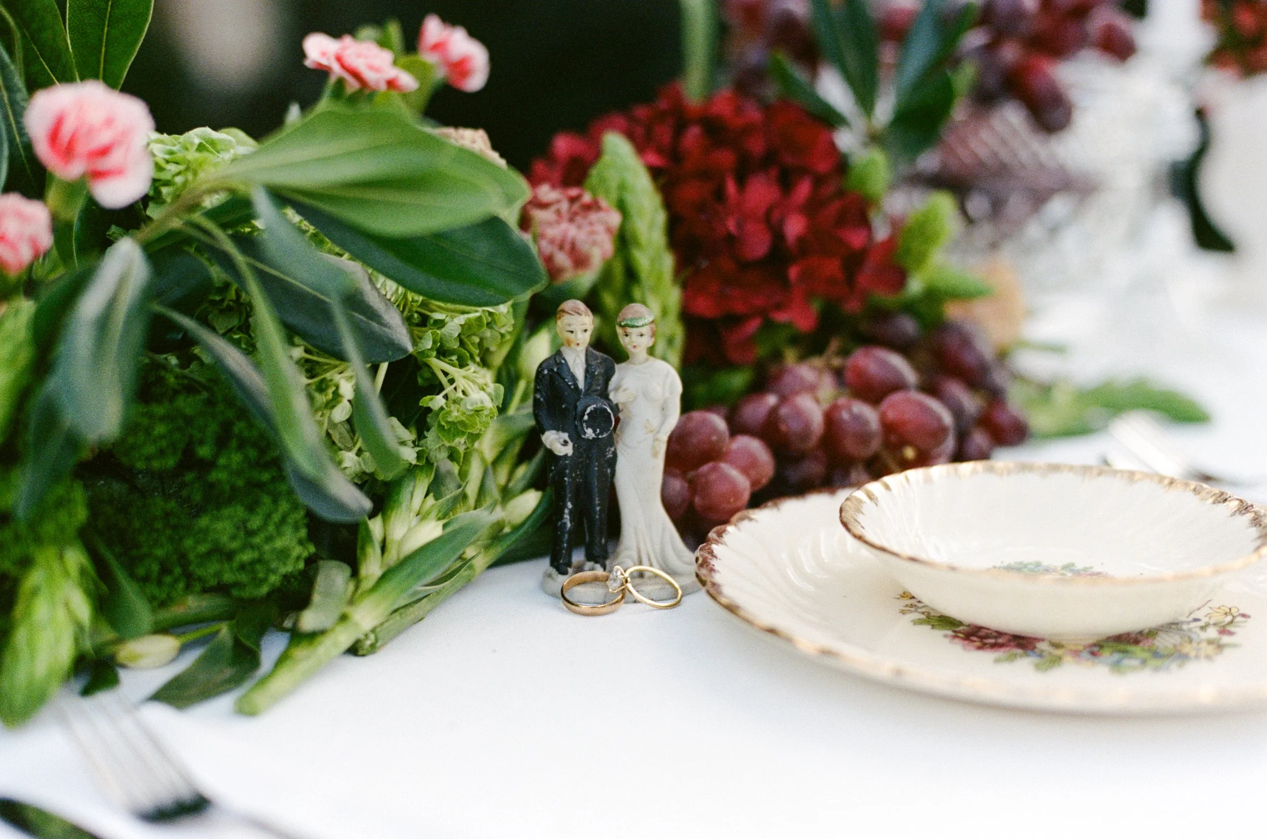 Wedding table centerpiece with flowers and miniature wedding figurines, wedding rings, a white ceramic plate with floral design, and a bunch of grapes in the background.