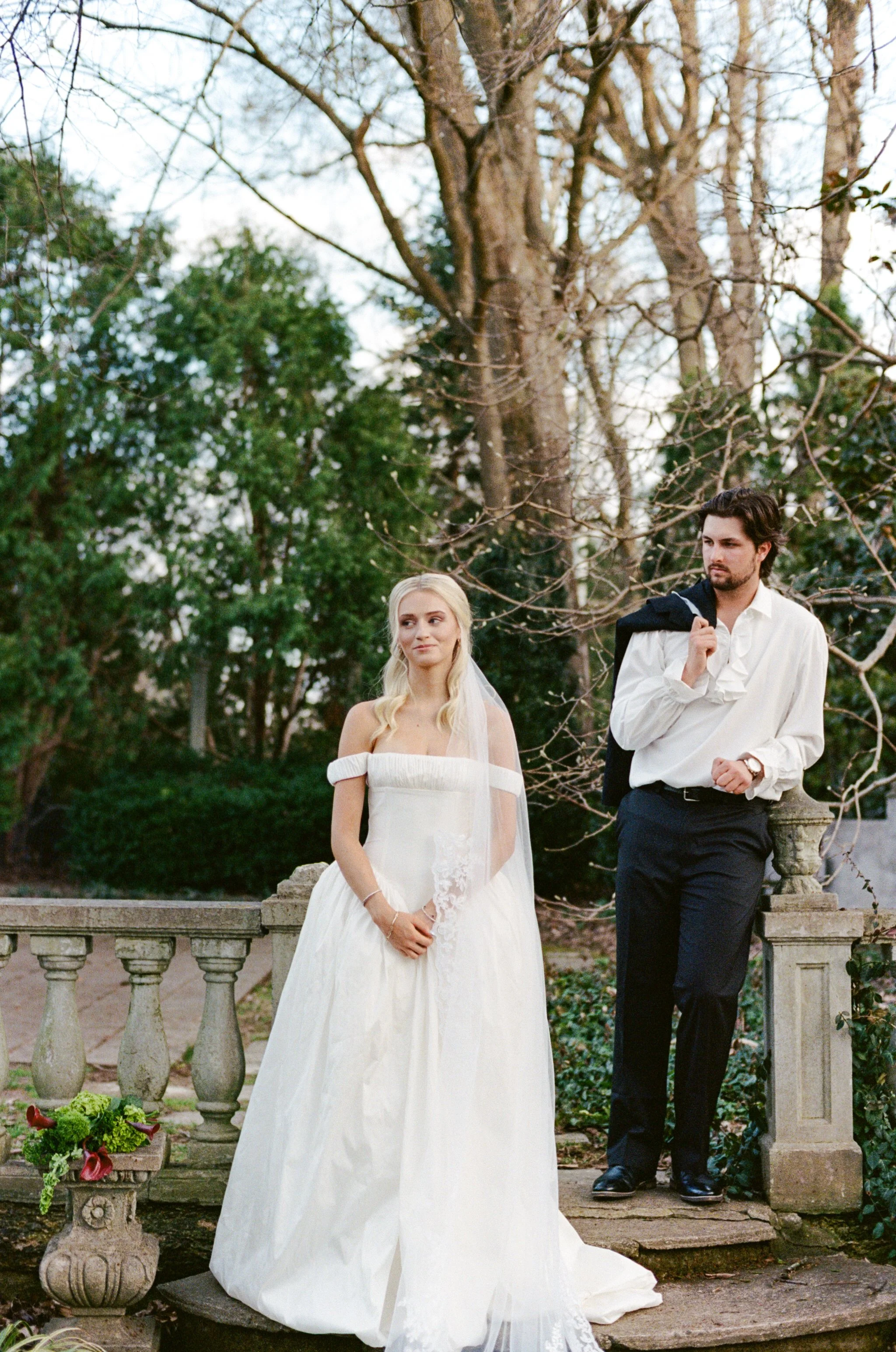 A bride and groom standing outdoors by a stone railing, with trees and greenery in the background. The bride is wearing a white wedding gown with off-the-shoulder sleeves and a veil. The groom is dressed in a white shirt and black pants, holding a ja