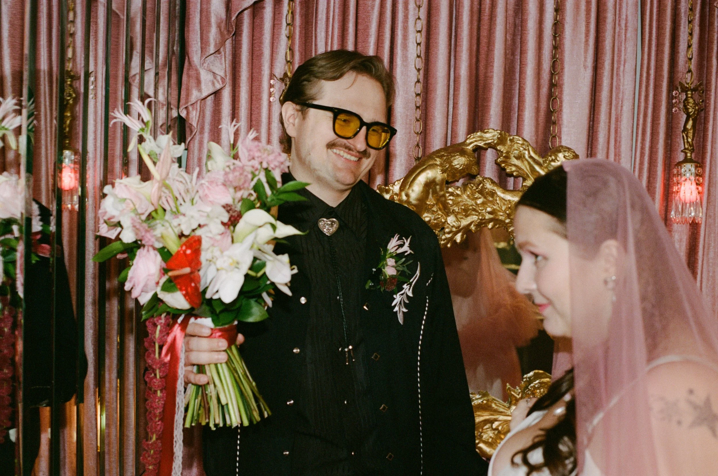 A man with brown hair, mustache, and sunglasses smiling while holding a bouquet of pink and white flowers at a celebratory event with pink curtains and gold decorative elements in the background. A woman with dark hair is partially visible in the for