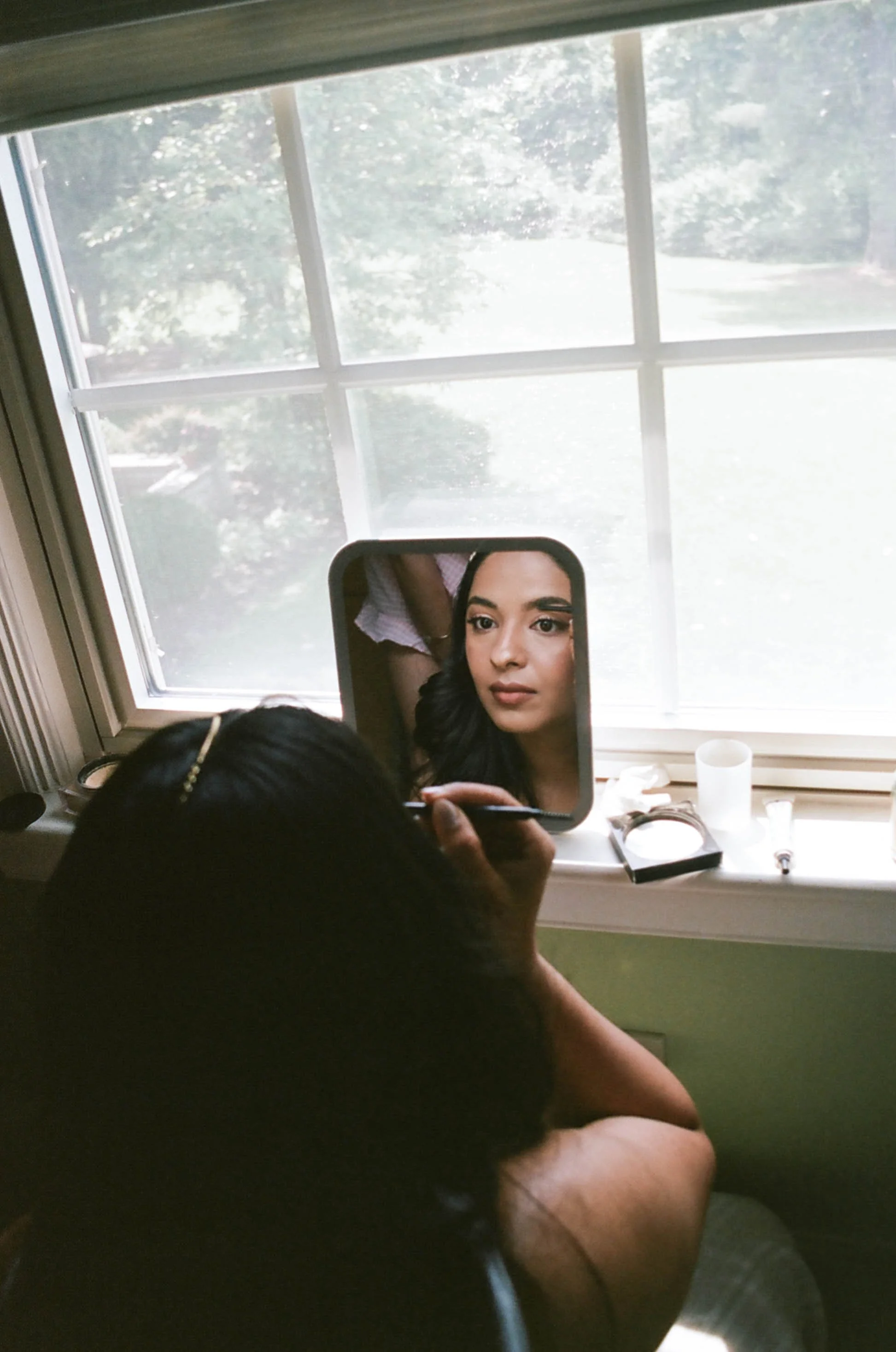 A person with dark hair is holding a mirror, taking a selfie in a room with a window showing trees outside. The person's face is reflected in the mirror, and they are centered in the image.