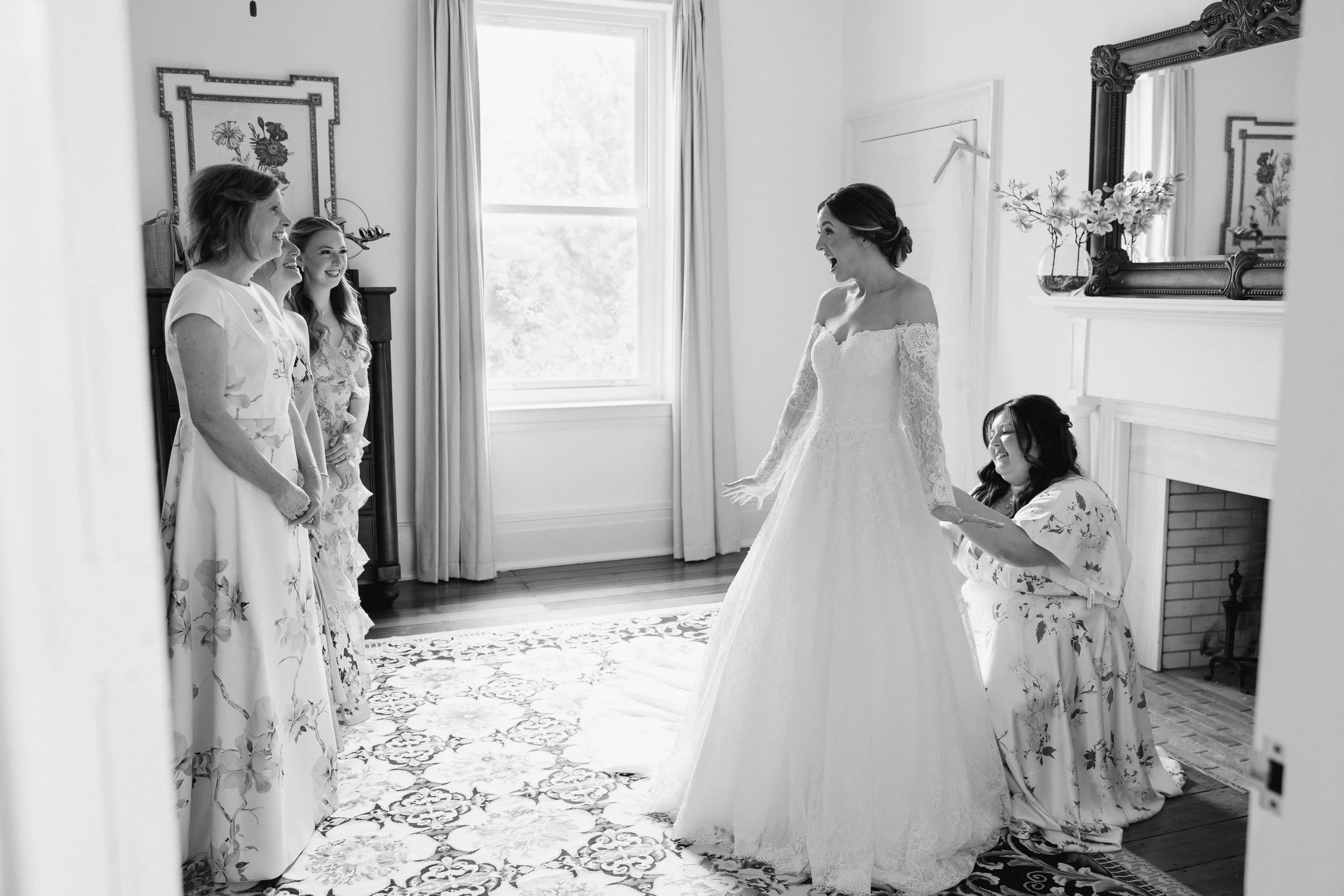 A bride in a lace wedding dress smiling excitedly at her bridesmaids in floral robes in a well-lit room with a window, picture frames, and a mirror.